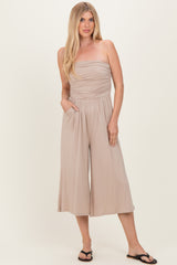 woman wearing a beige strapless jumpsuit with a ruched bodice and side pockets, standing and smiling in flip flops