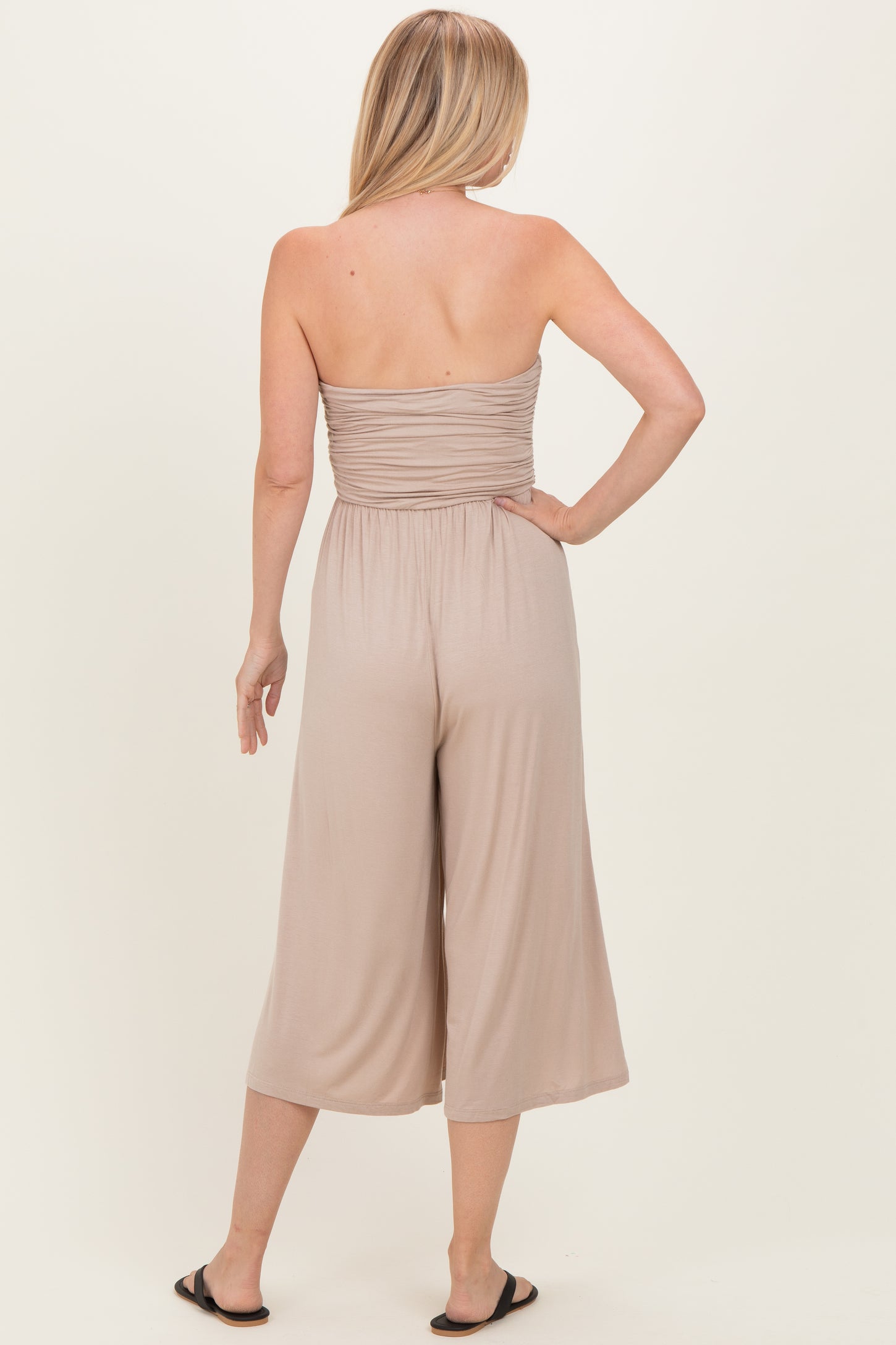 woman wearing a taupe strapless jumpsuit with a gathered bodice and wide leg cropped pants, viewed from the back