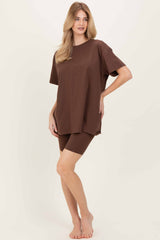 woman wearing a brown oversized t-shirt and matching knee-length shorts, standing barefoot against a light background