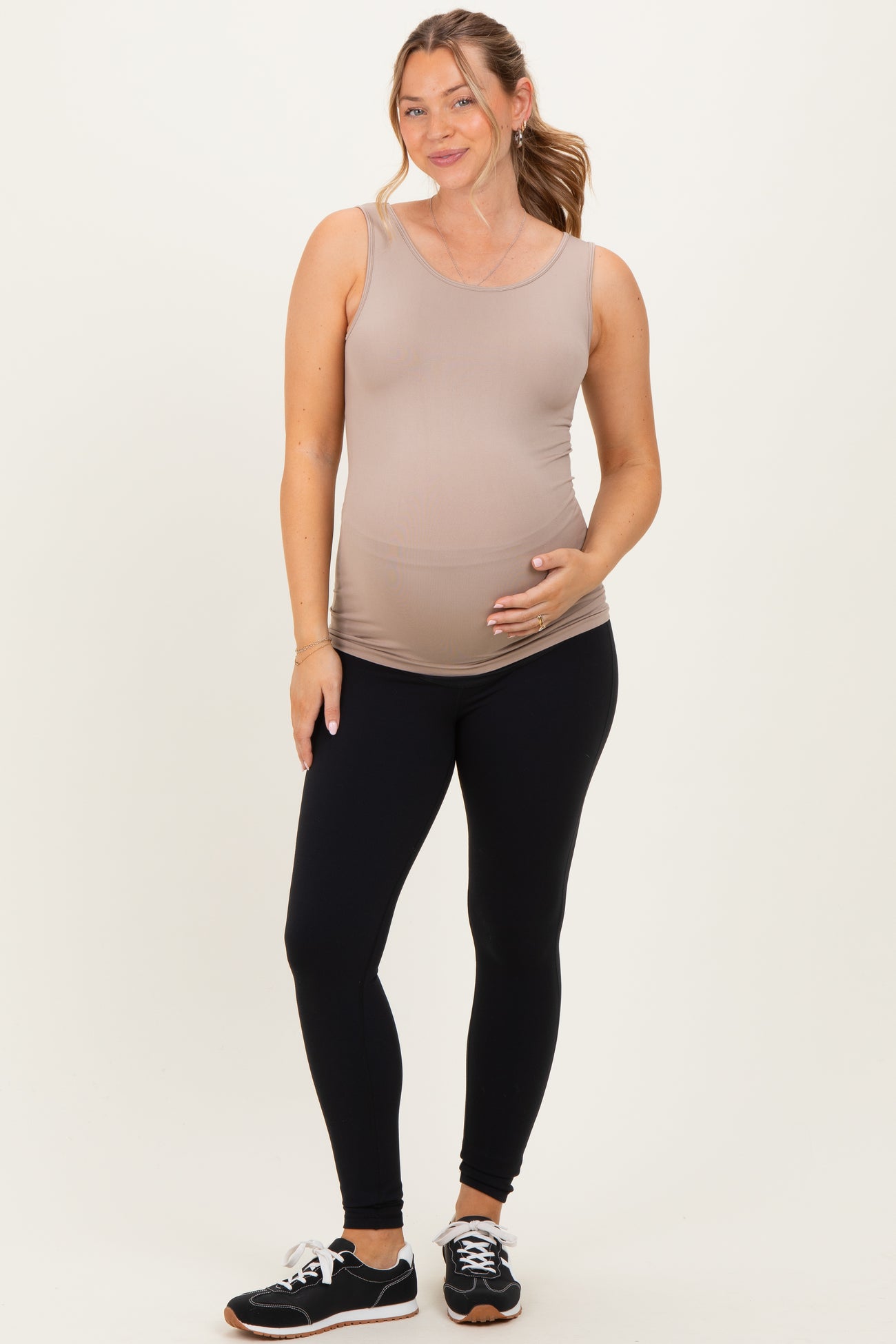 woman wearing a nude maternity tank top and black leggings, gently holding her baby bump