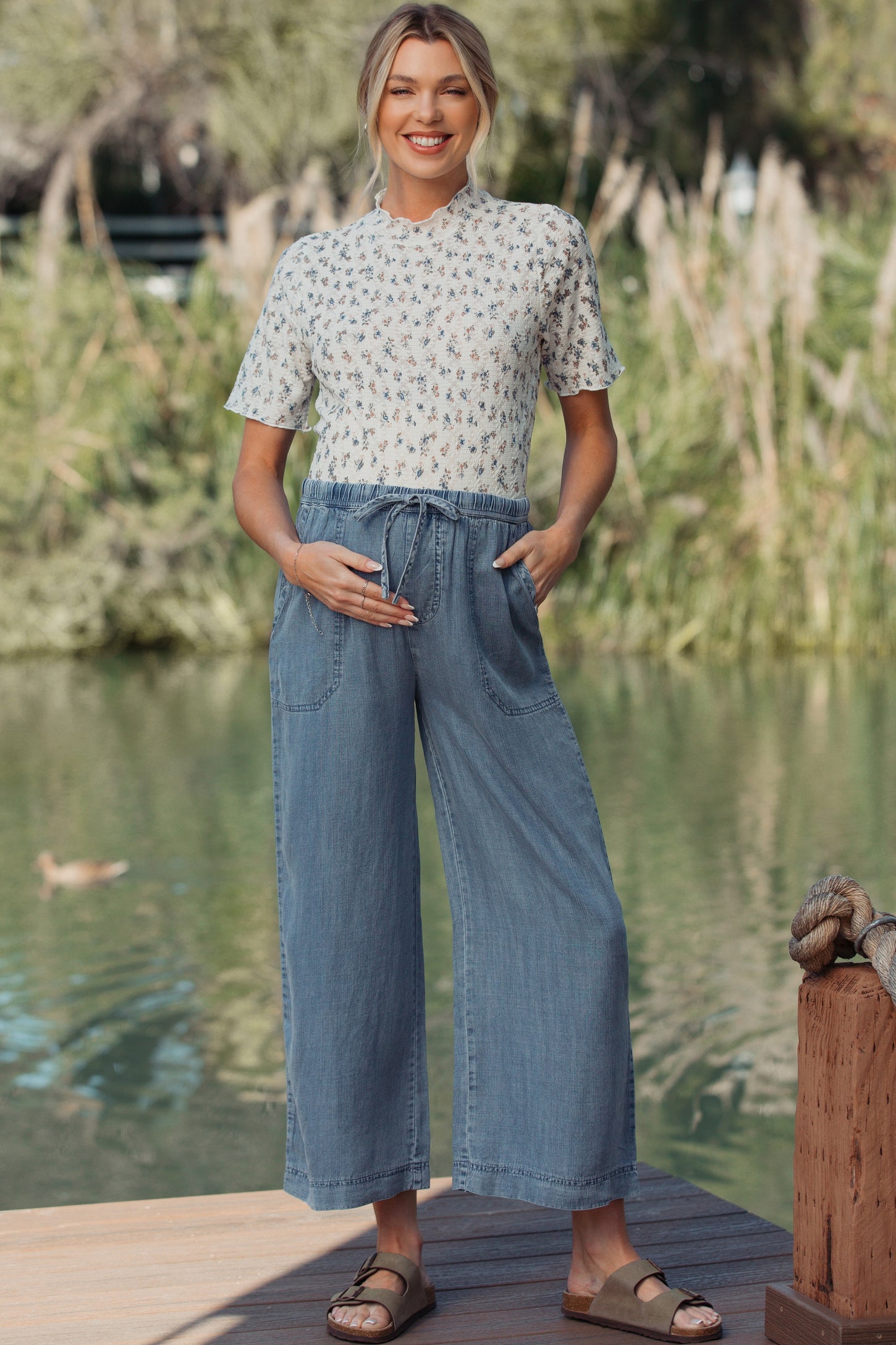woman wearing a floral print maternity top and denim wide-leg pants, holding her baby bump outdoors by a lake