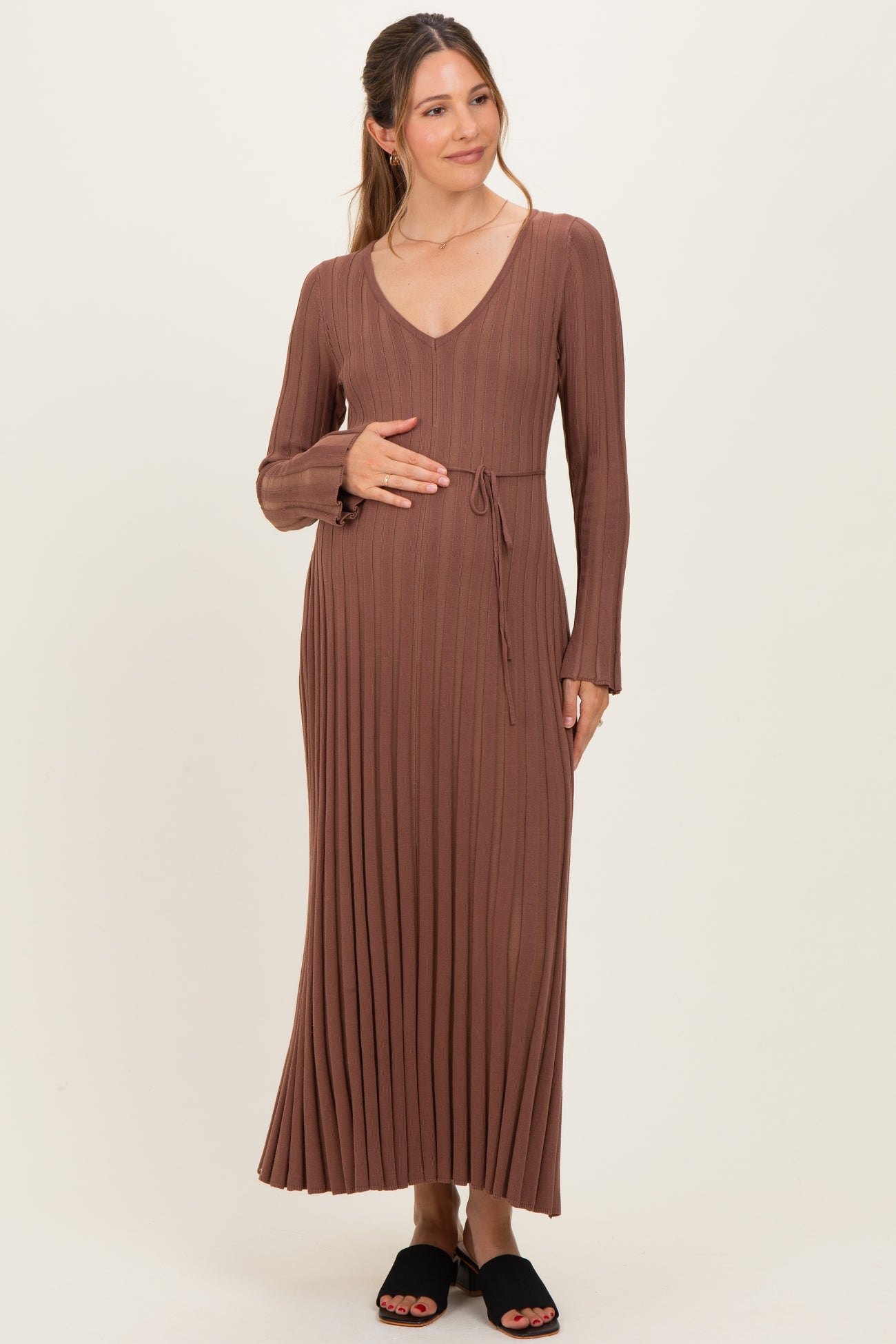 woman wearing a long brown ribbed maternity dress with a v-neck and tie waist, gently cradling her baby bump