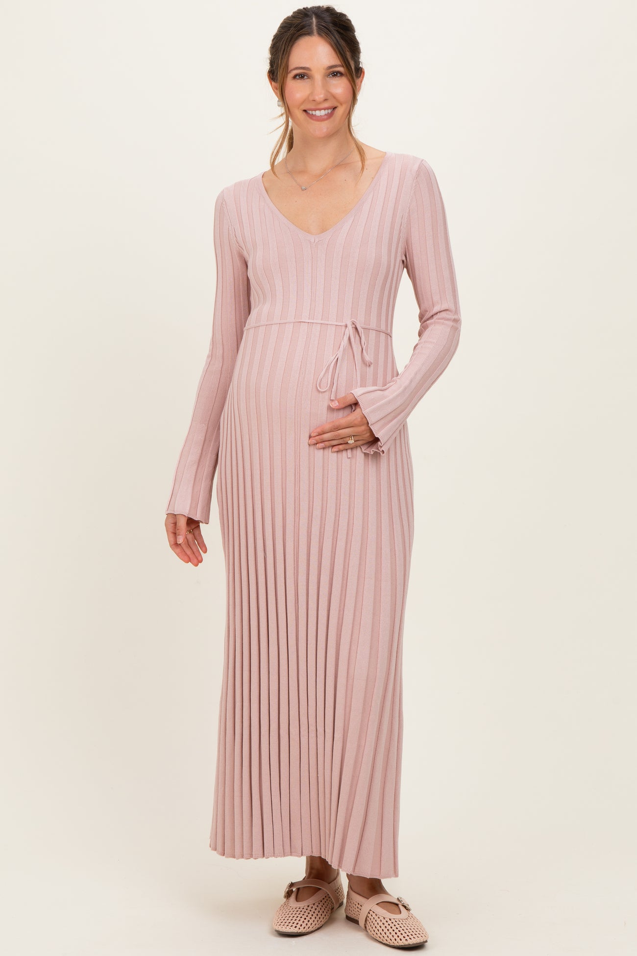 woman wearing a light pink ribbed maternity dress with long sleeves and a tie waist, holding her baby bump