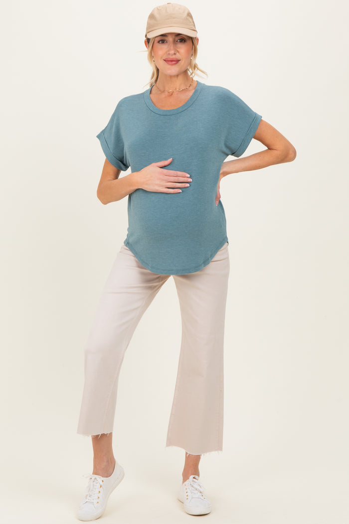 woman wearing a teal maternity top and light beige cropped pants, holding her baby bump