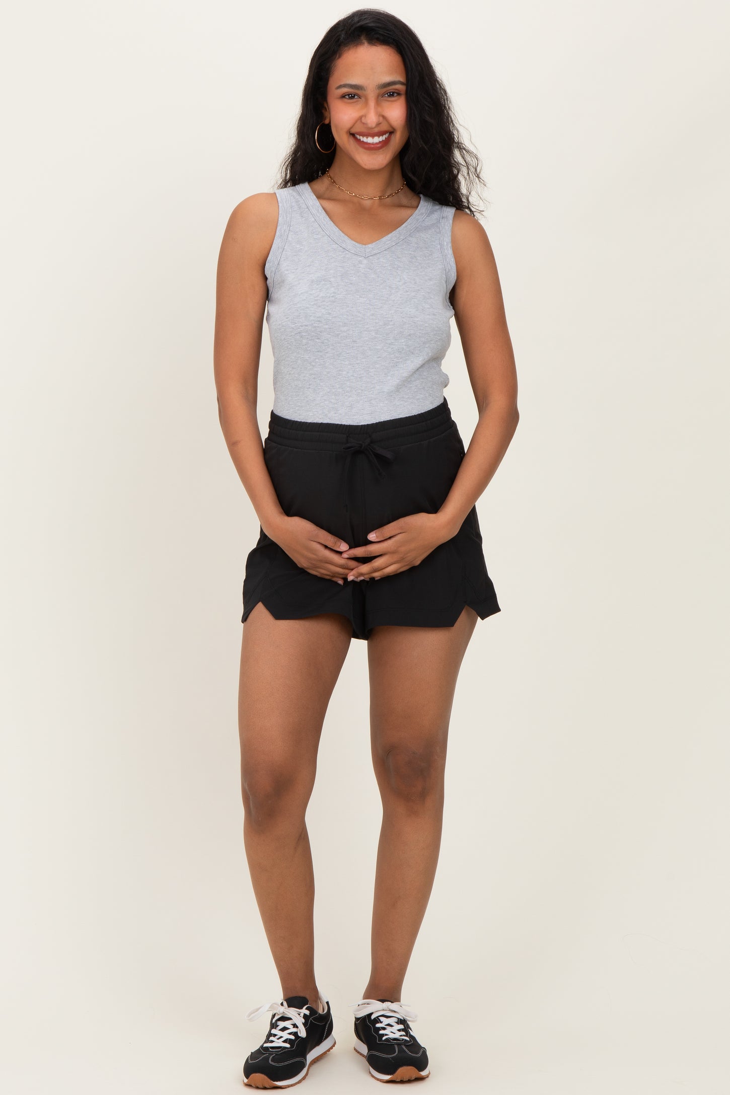 woman wearing a gray sleeveless top and black maternity shorts, holding her baby bump
