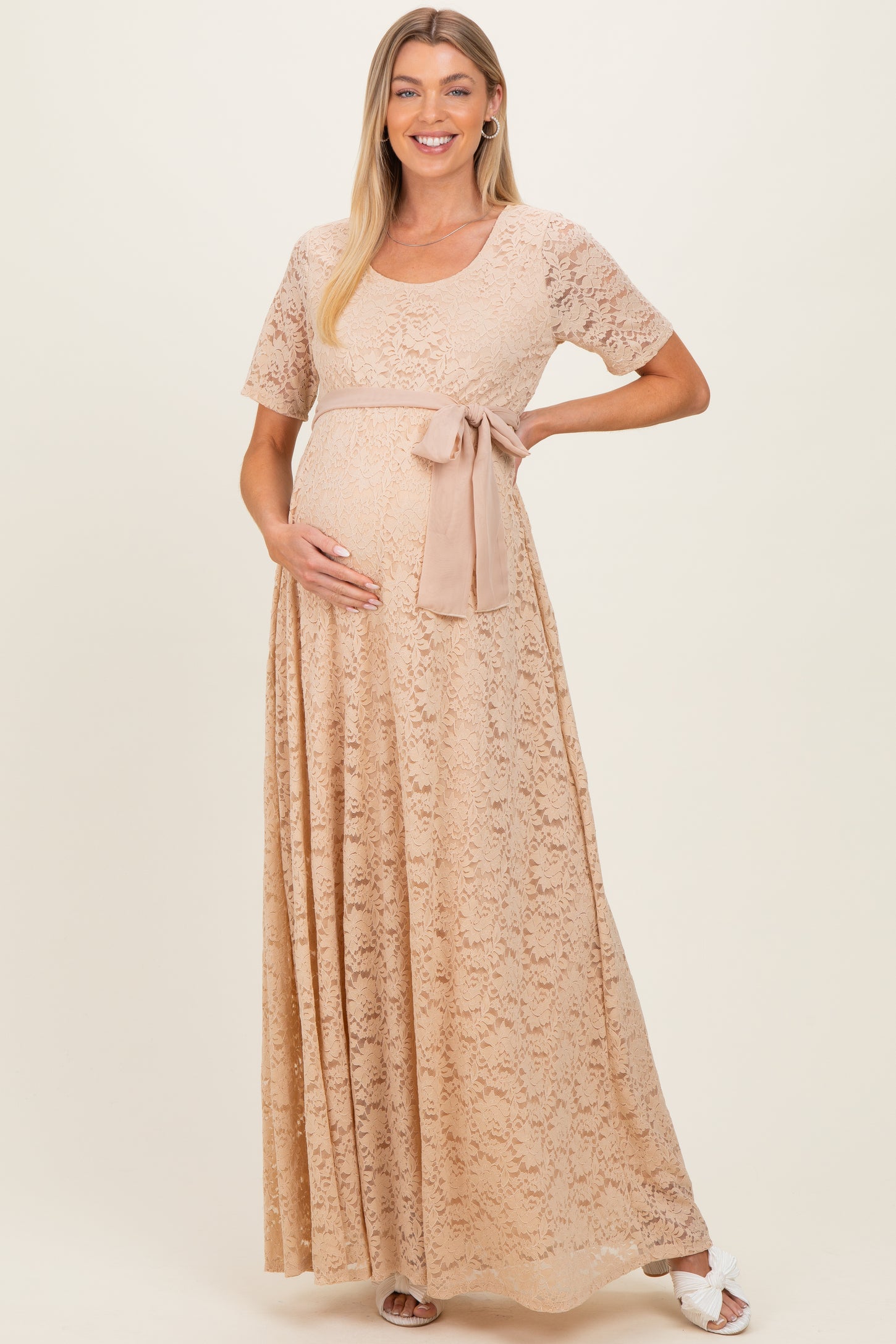 woman wearing a beige lace maternity maxi dress with a tied waist and holding her baby bump