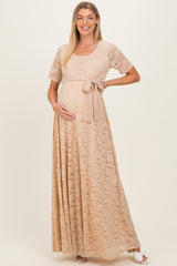 woman wearing a beige lace maternity maxi dress with a tied waist and holding her baby bump