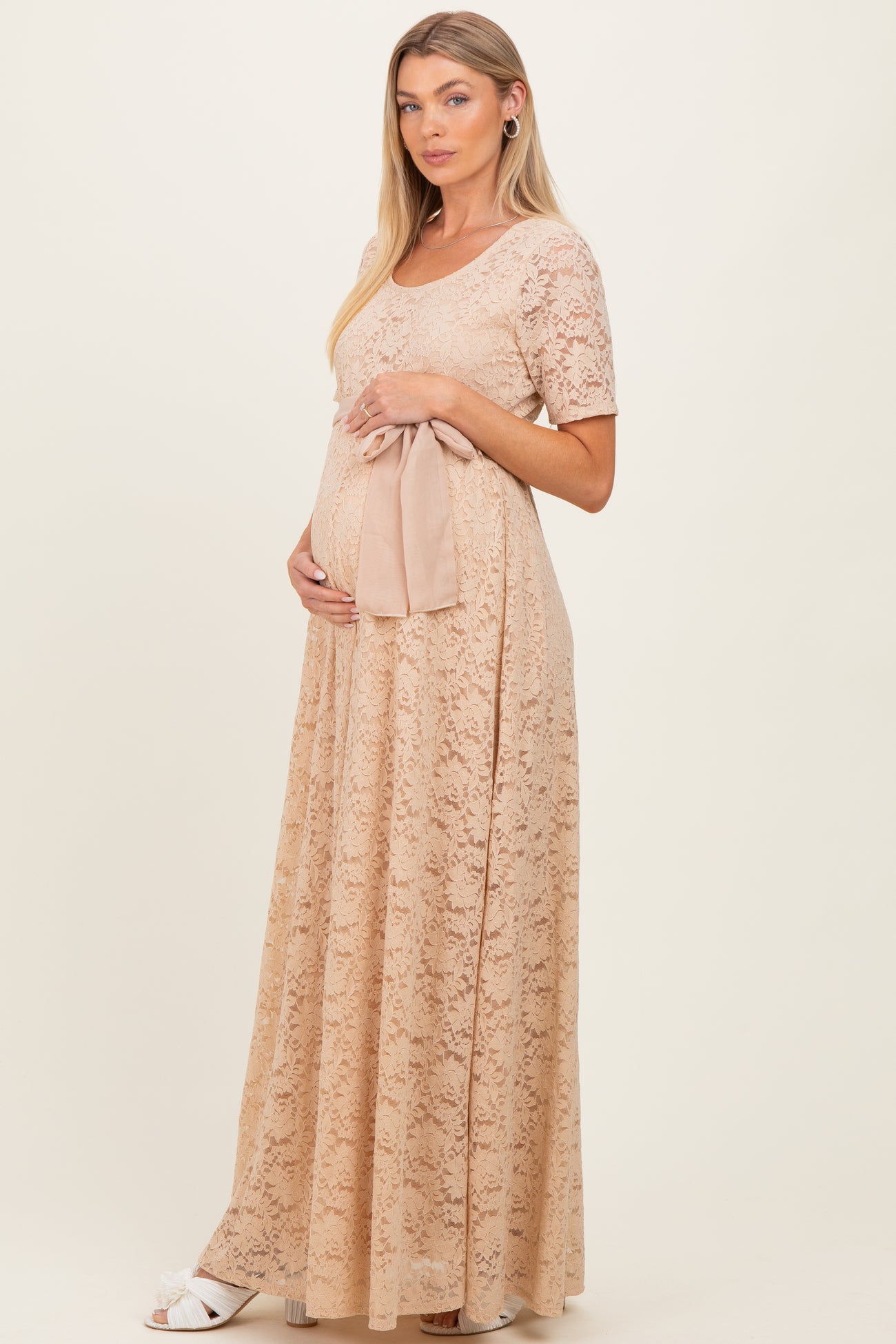 woman wearing a beige lace maternity gown with short sleeves, gently holding her baby bump