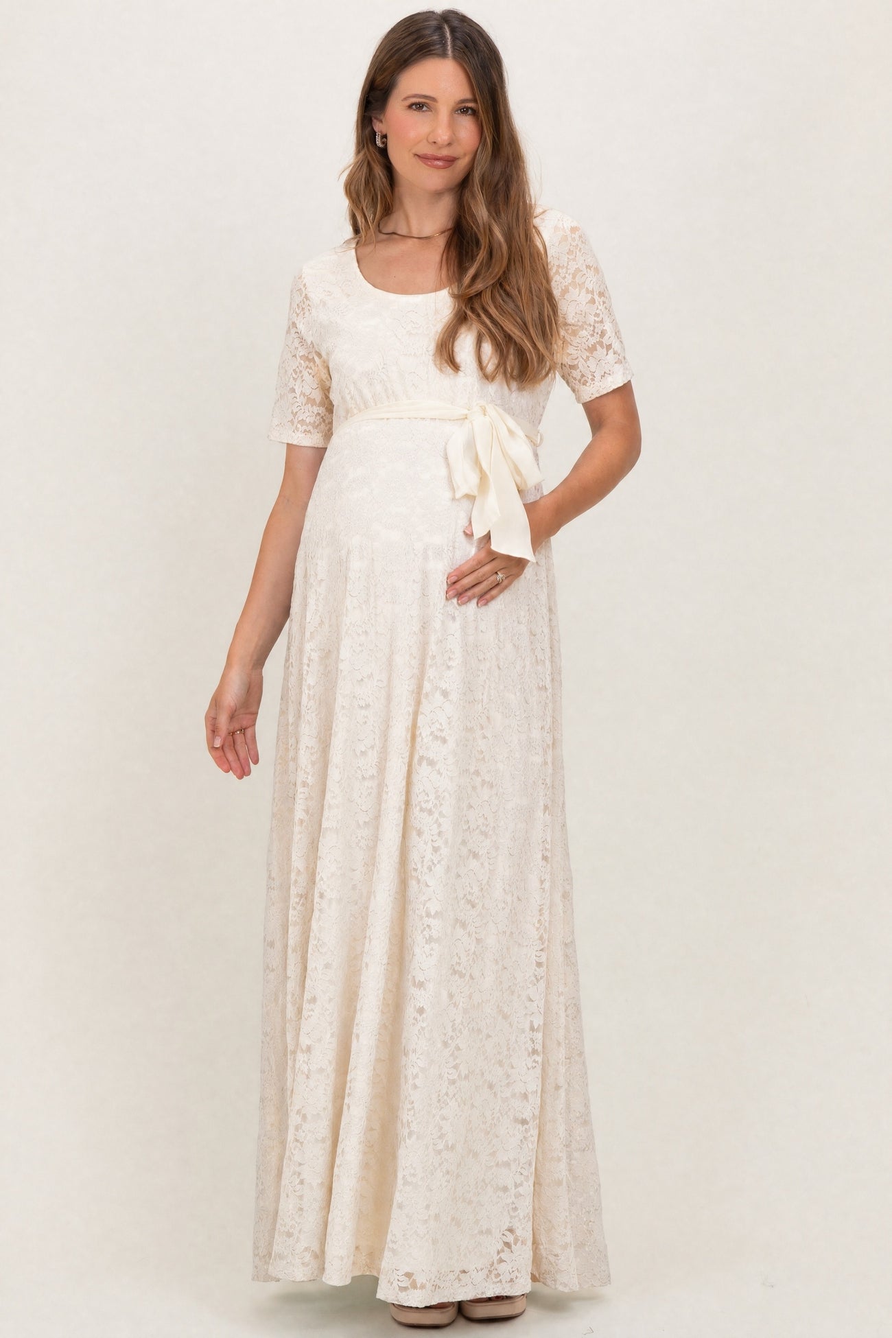 woman wearing a lace cream maternity maxi dress with short sleeves and a bow detail at the waist, showing a baby bump