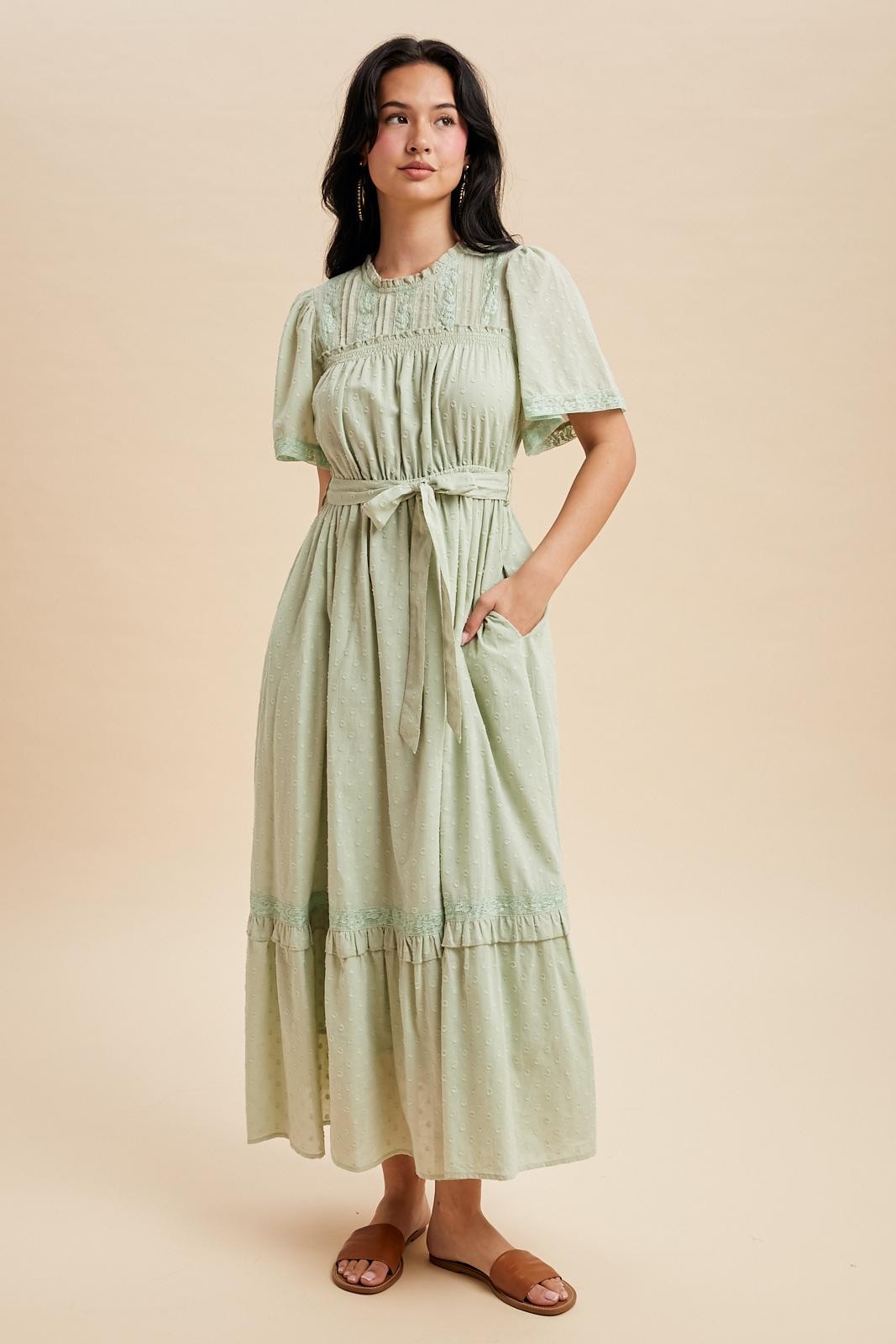 woman wearing a light green maxi dress with short sleeves, a belted waist, and pockets, standing against a neutral background