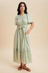 woman wearing a light green maxi dress with short sleeves, a belted waist, and pockets, standing against a neutral background
