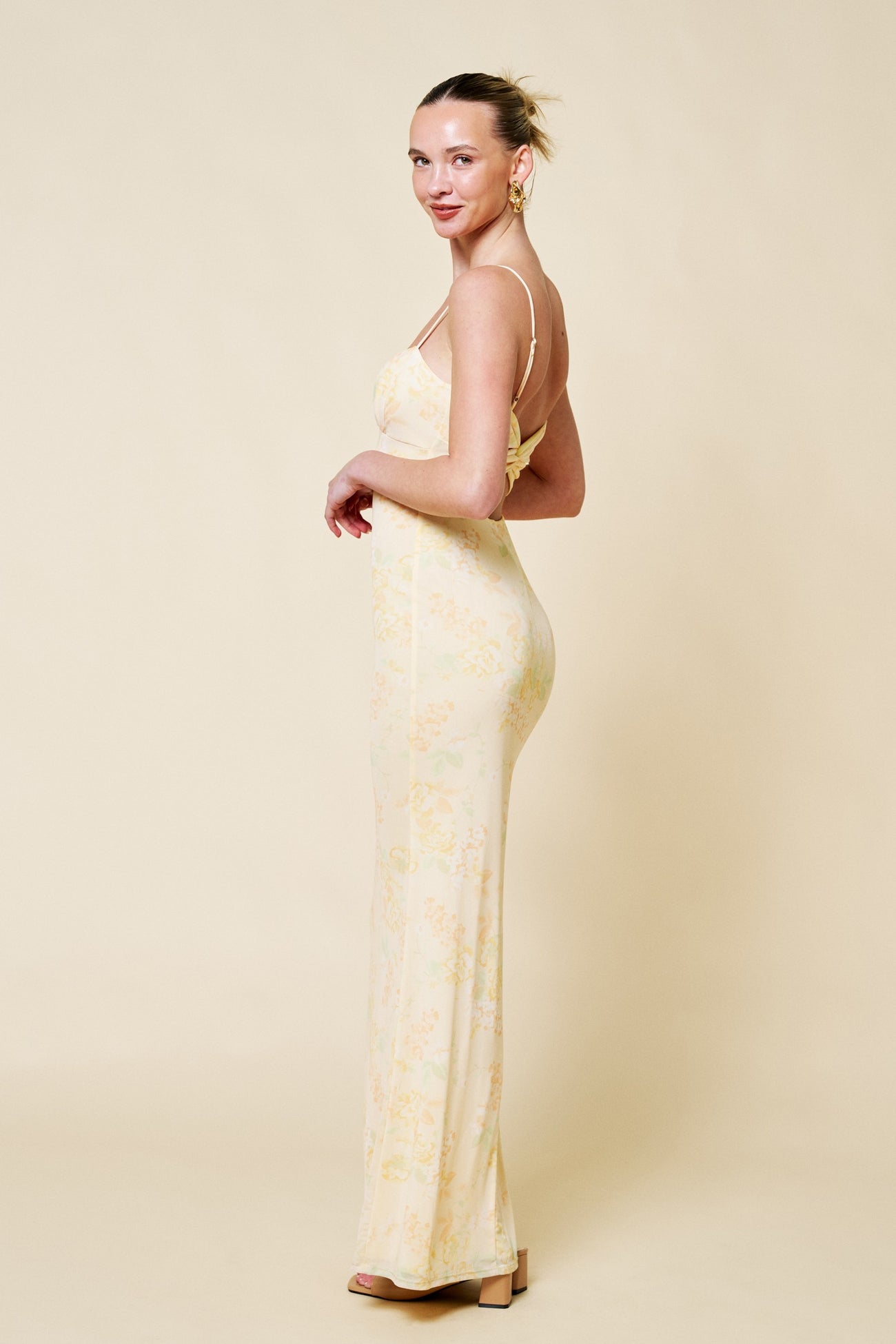 woman wearing a yellow floral spaghetti strap maxi dress, standing sideways with a soft smile and hair styled up