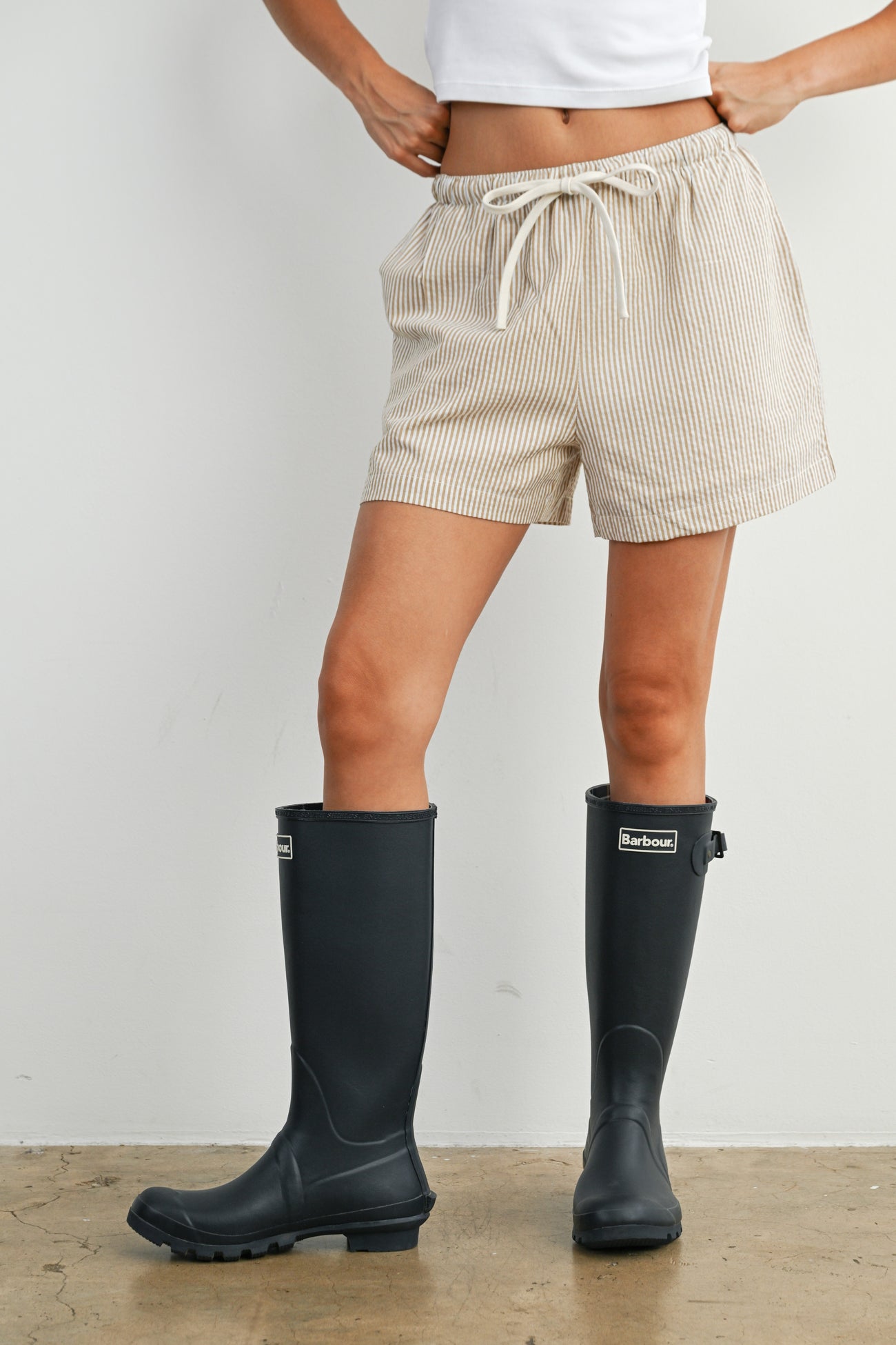 woman wearing beige striped shorts and black rubber rain boots, standing on a textured floor