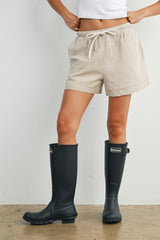 woman wearing beige striped shorts and black rubber rain boots, standing on a textured floor