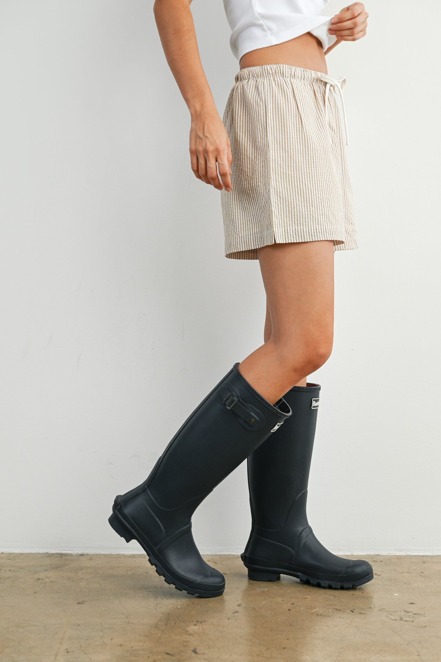 woman wearing a white t-shirt, striped shorts, and black rain boots standing against a wall