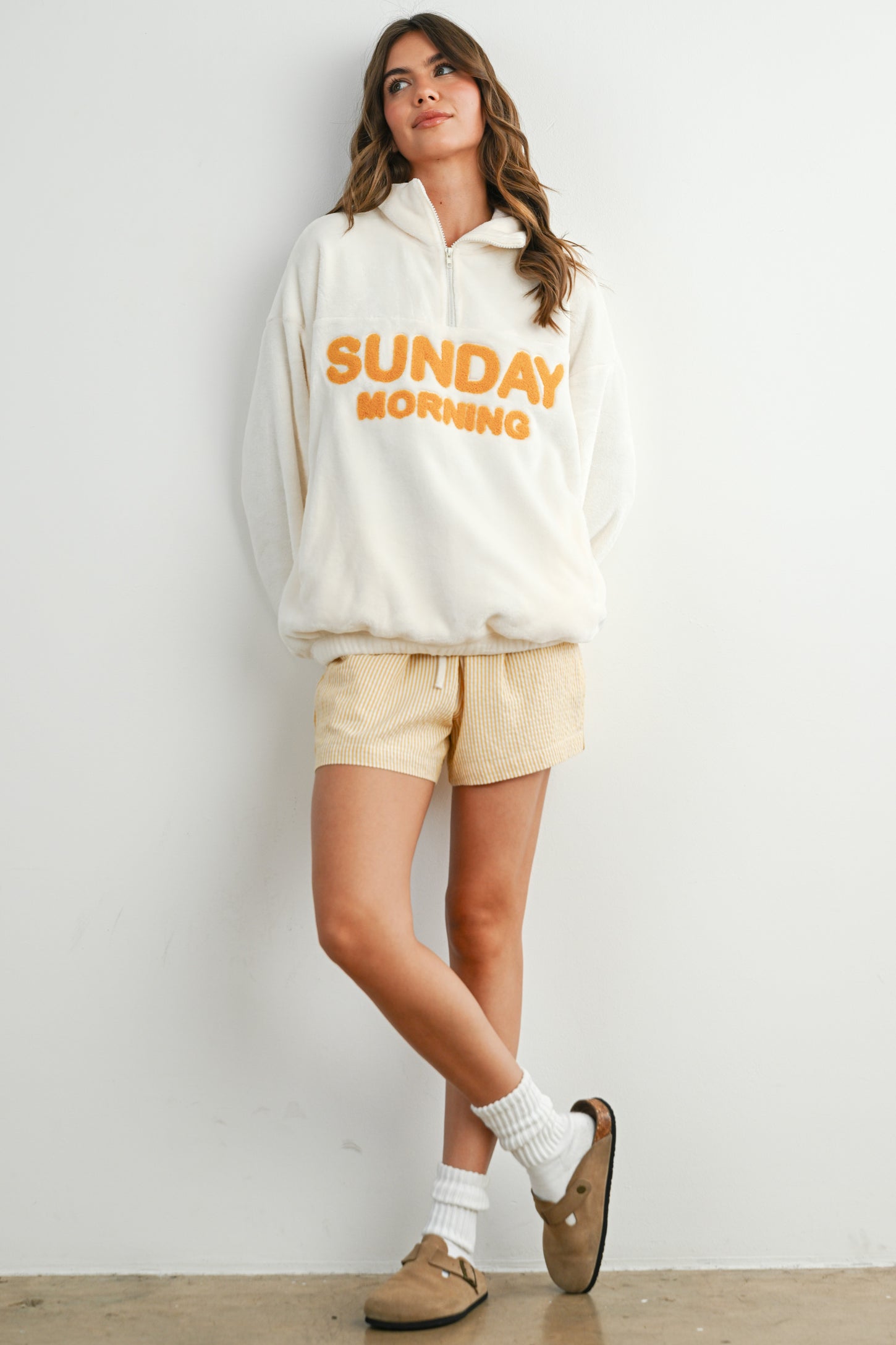 woman wearing a cream fleece pullover with "SUNDAY MORNING" text and striped shorts, styled with socks and slippers