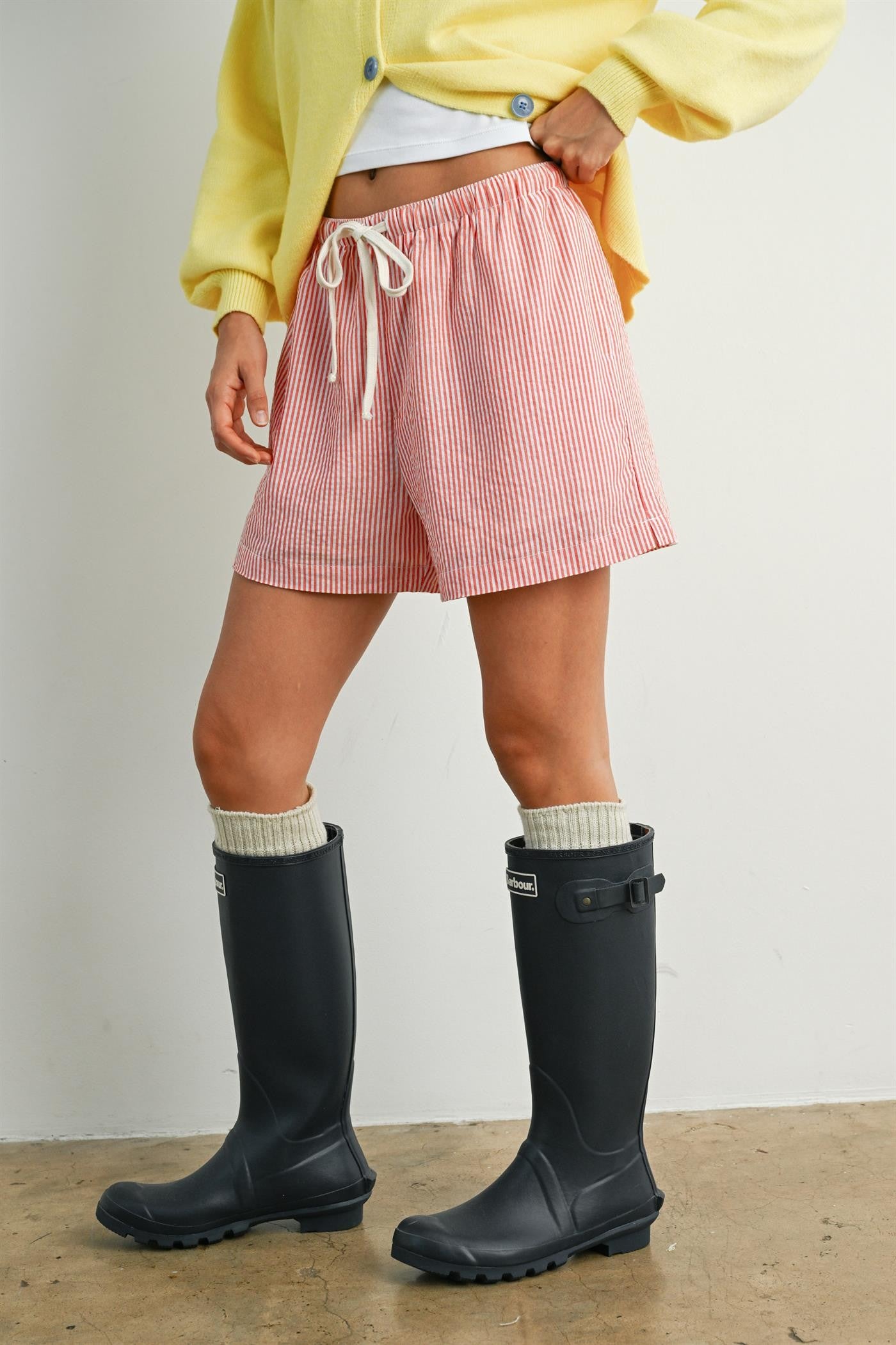 woman wearing red and white striped shorts with a drawstring and black rubber boots, styled with a yellow cardigan