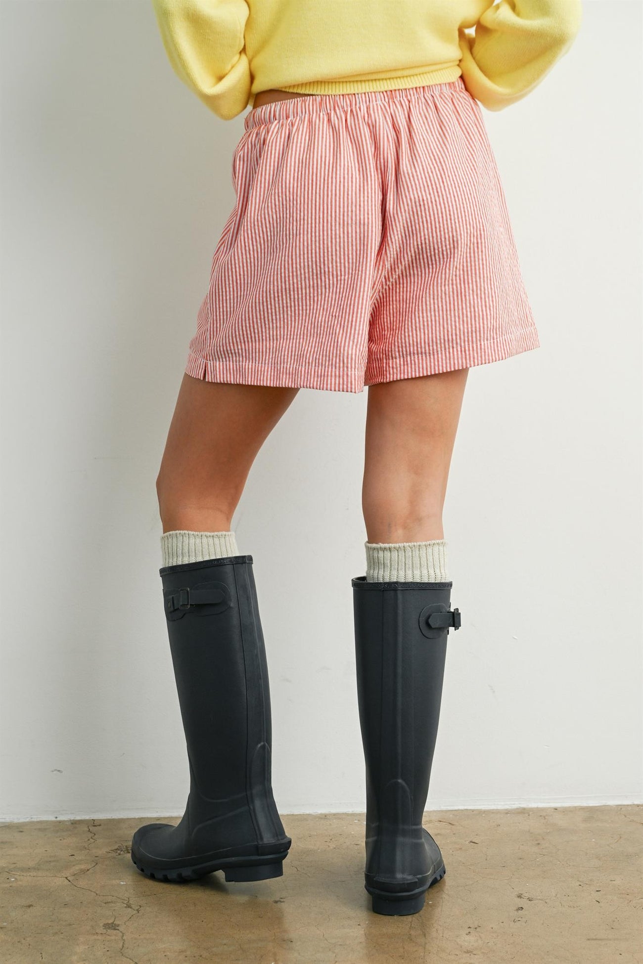woman wearing red and white striped shorts and tall black rubber boots with gray knit socks, viewed from behind