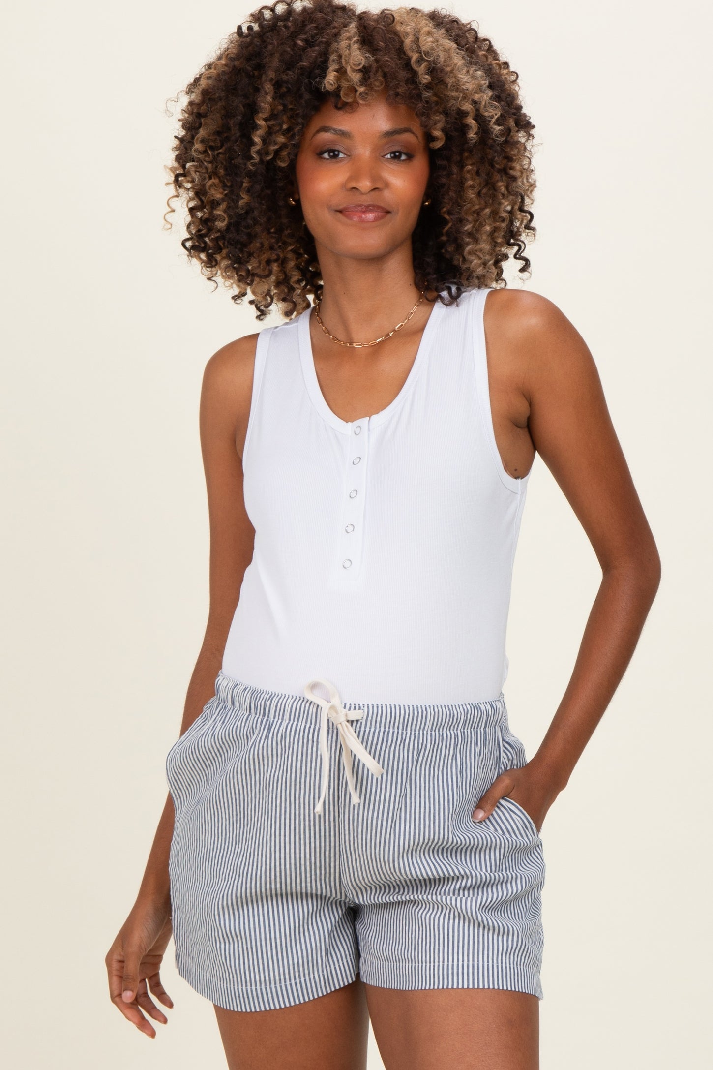 woman wearing a white maternity tank top with buttons and striped shorts, hands in pockets, showing her baby bump