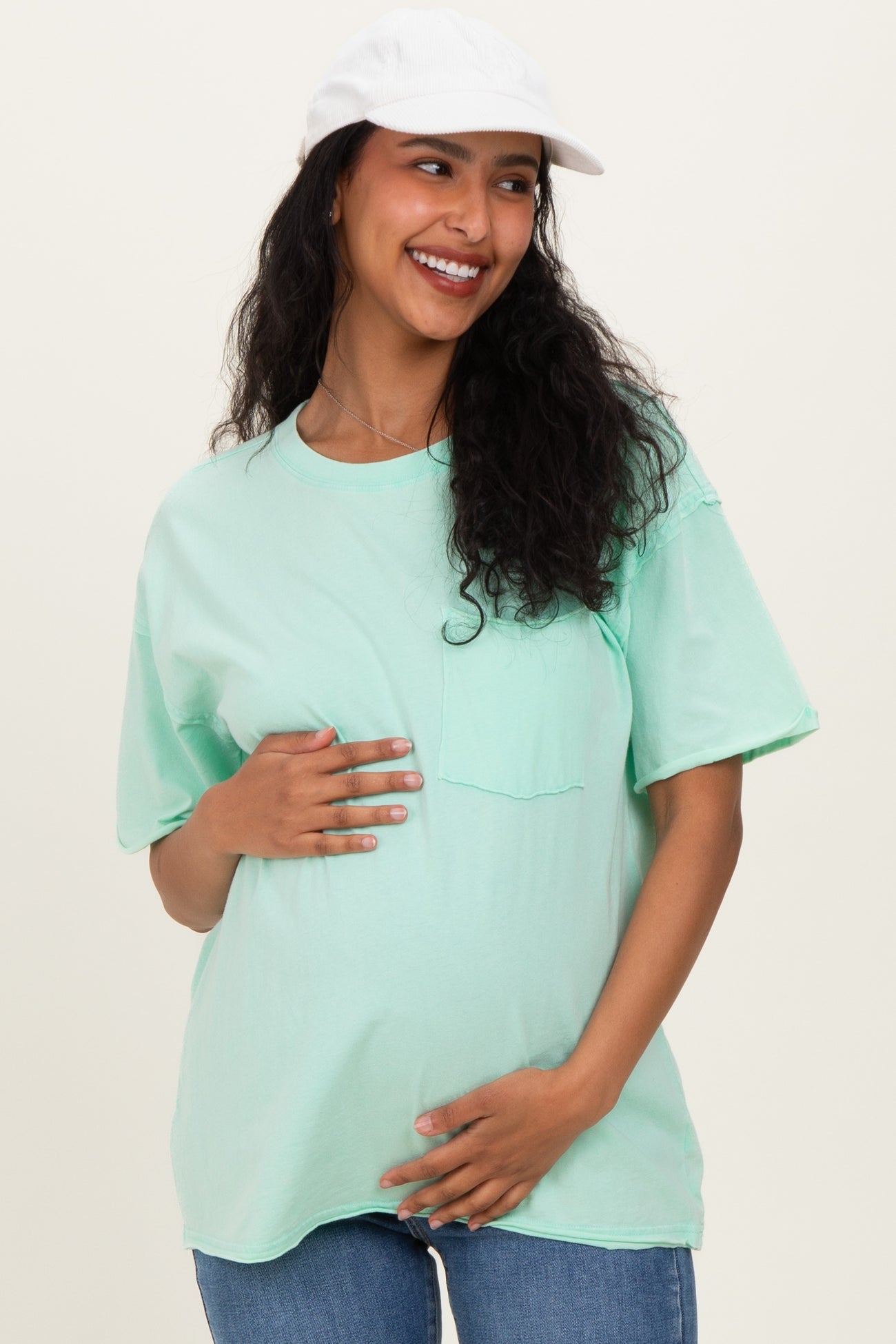 woman wearing a mint green oversized maternity t-shirt and holding her baby bump, smiling with a baseball cap on