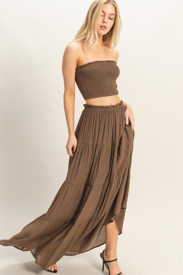 woman wearing a brown off-shoulder smocked crop top and a matching flowy maxi skirt, posing elegantly