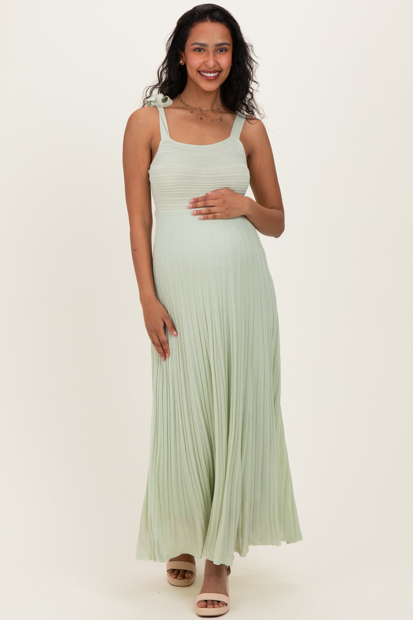 woman wearing a light green pleated maternity dress with a shoulder tie, holding her baby bump