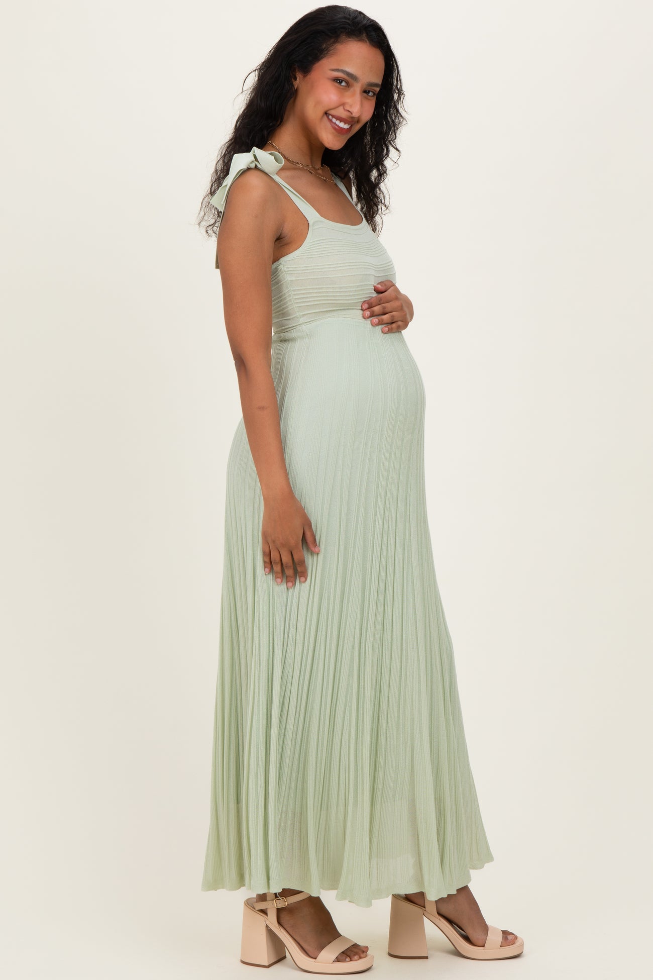 woman wearing a light green pleated maternity dress with a tie strap and holding her baby bump