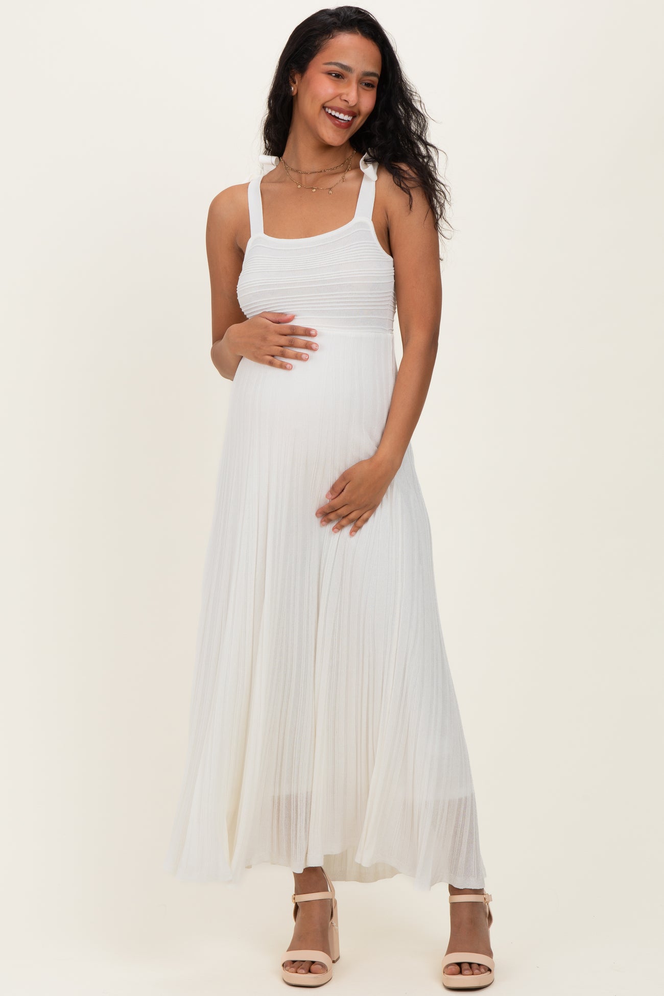 woman wearing a white pleated maternity dress with a fitted bodice and holding her baby bump