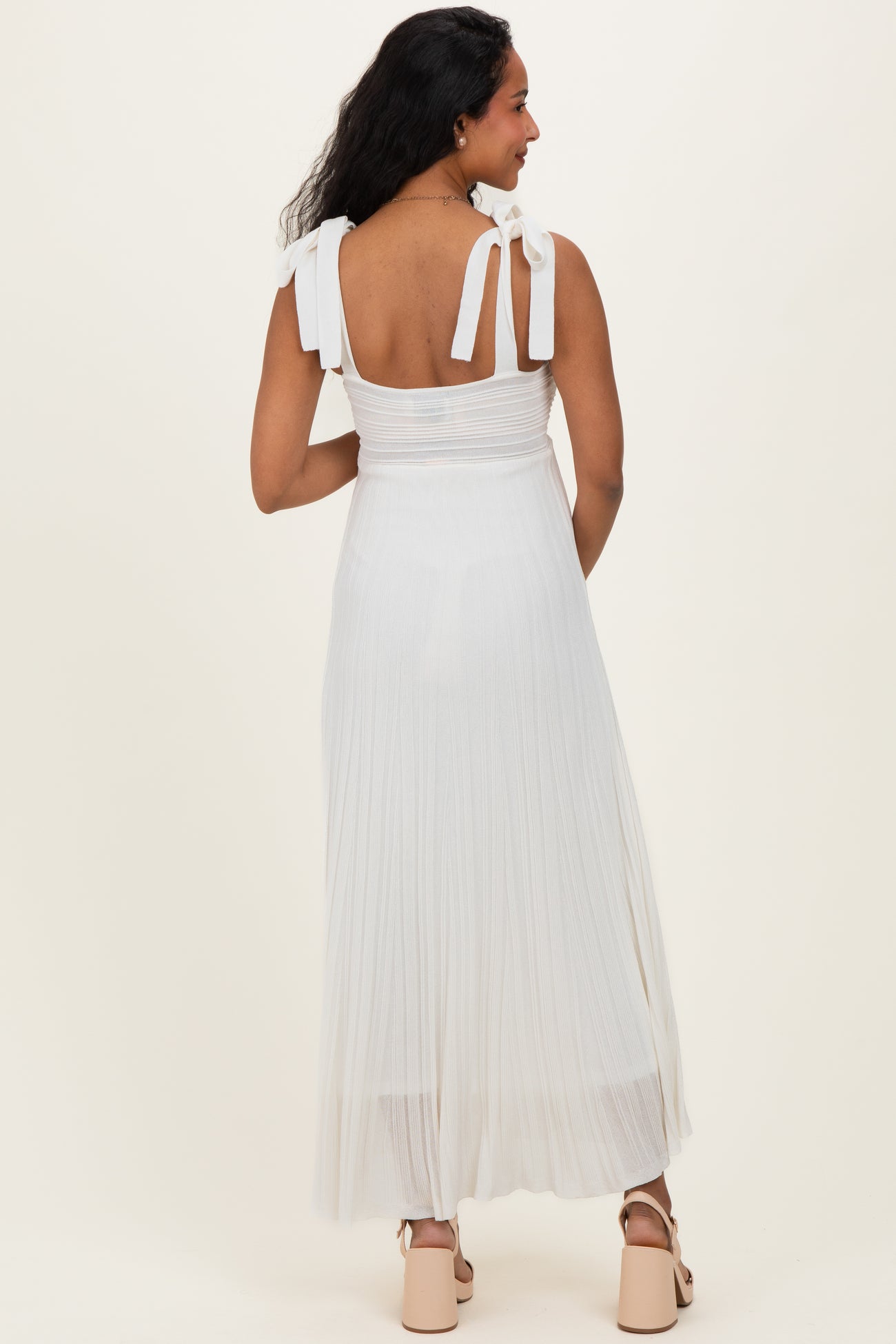 woman wearing a white pleated maternity dress with tie shoulder straps, showing her back and flowing skirt