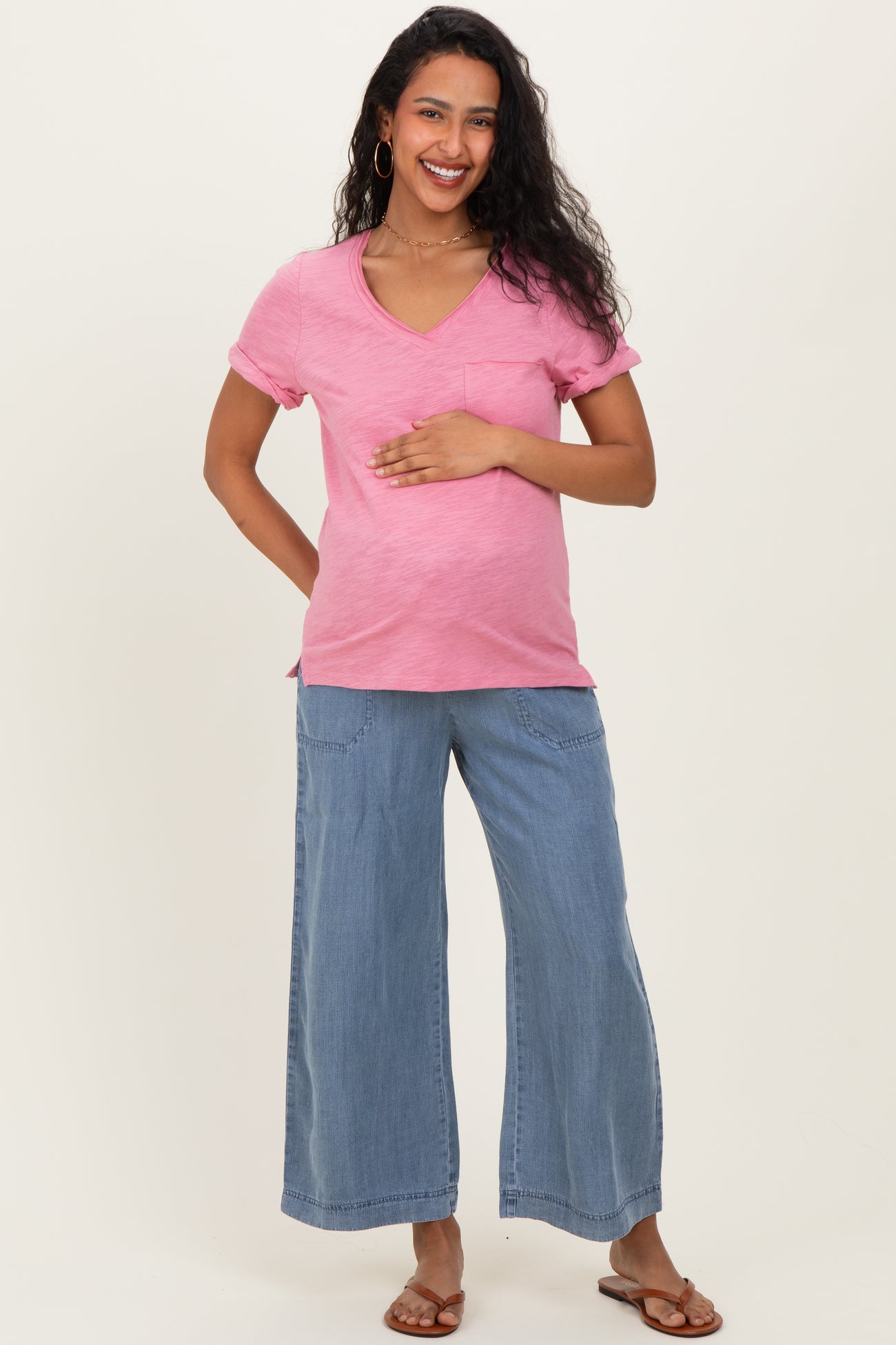 woman wearing a pink short sleeve maternity top and blue wide leg jeans, gently cradling her baby bump