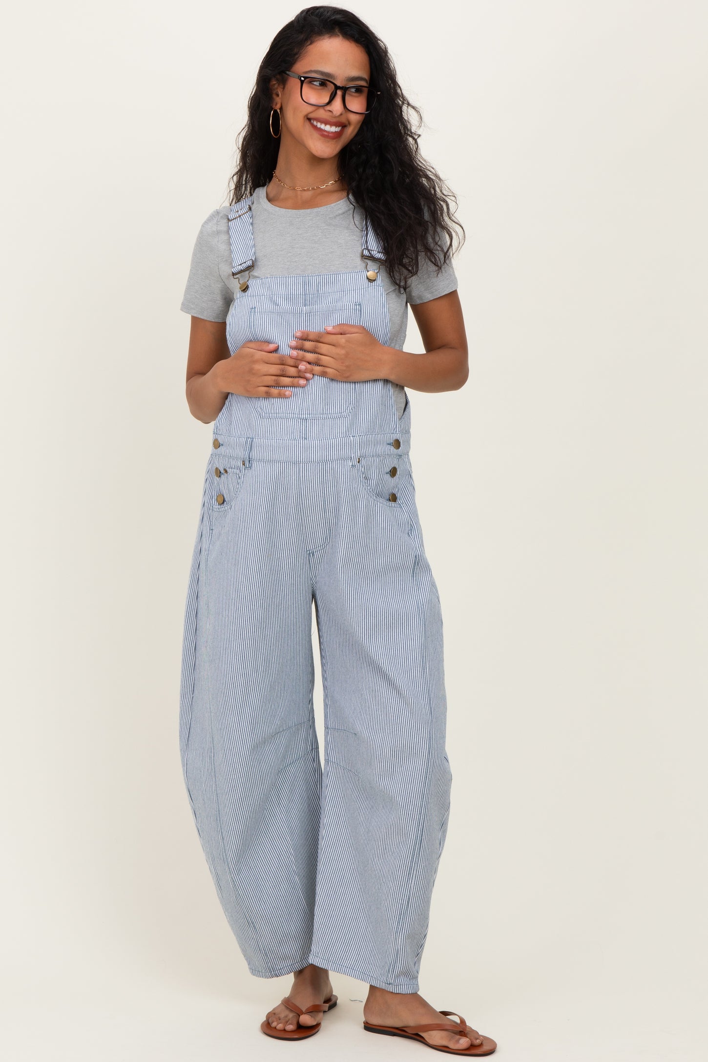 woman wearing a gray short sleeve shirt with blue and white striped maternity overalls and brown sandals