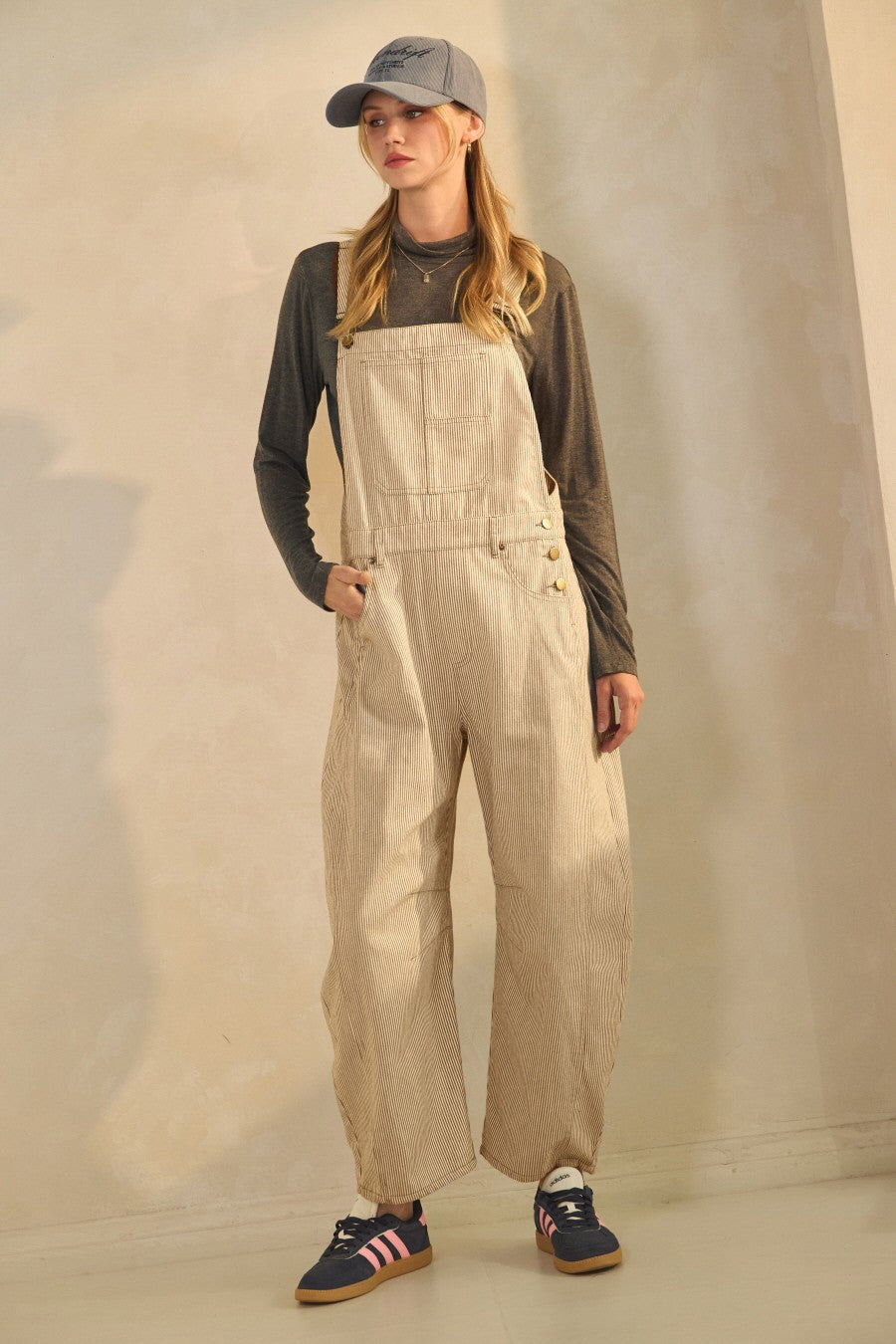 woman wearing beige striped overalls with a gray long sleeve top and a baseball cap, paired with navy sneakers