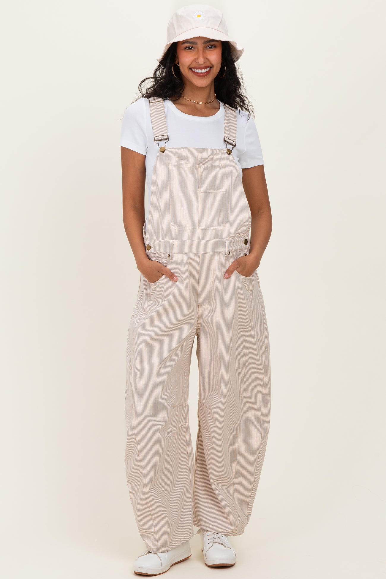 woman wearing a striped beige maternity overall with wide legs and a white t-shirt, smiling and posing with hands in pockets