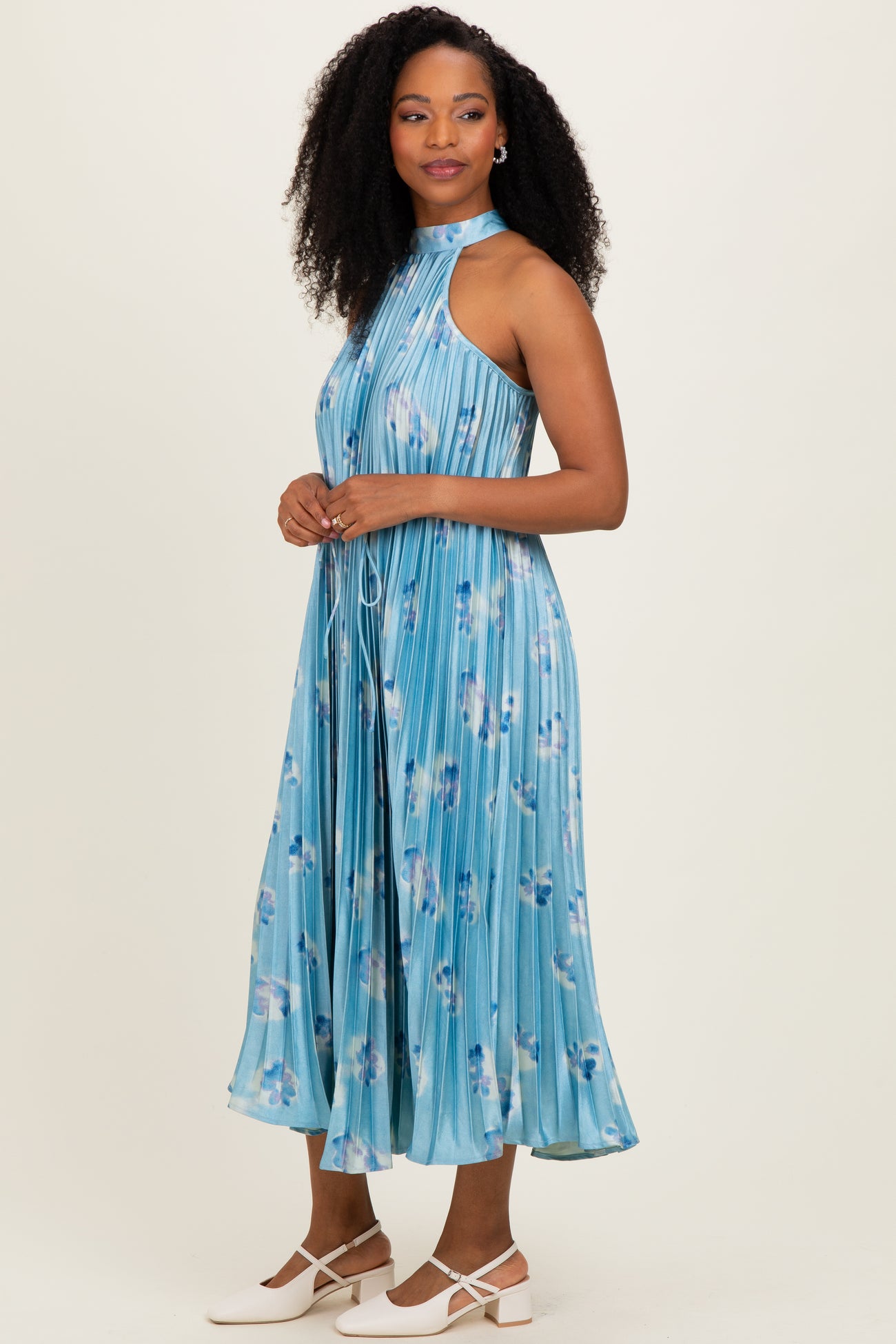 woman wearing a blue pleated midi dress with floral patterns and a halter neck, styled with white flats