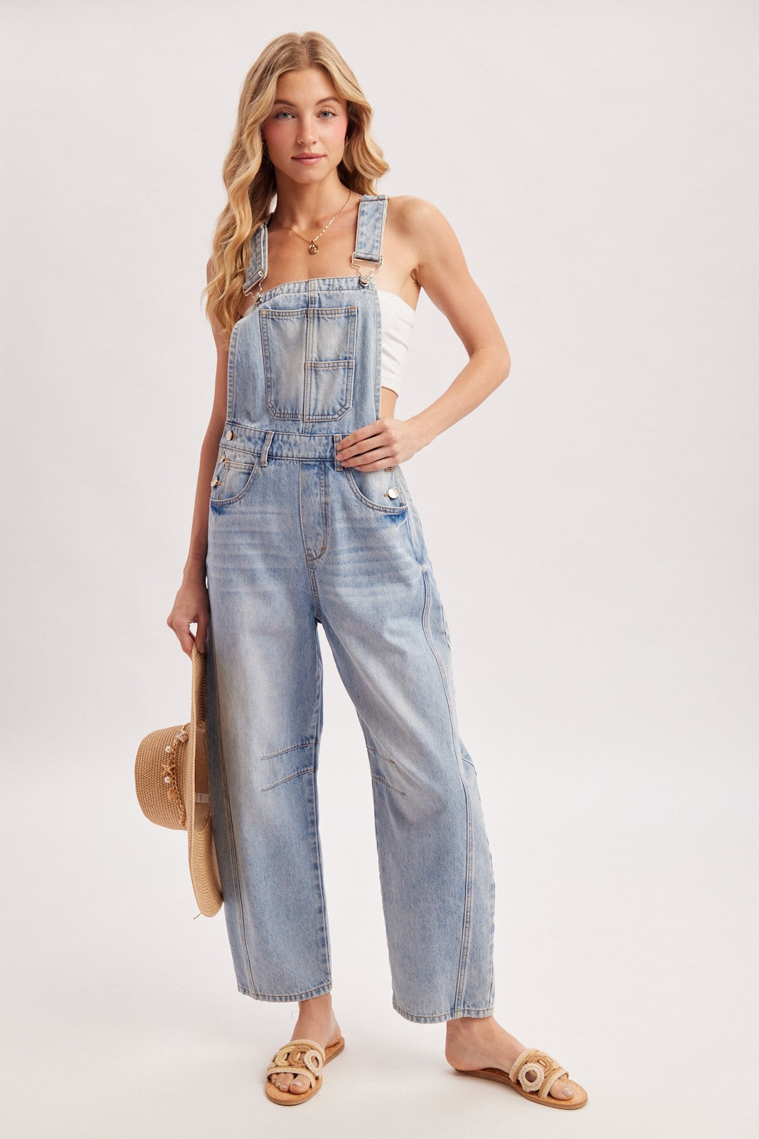 woman wearing a light blue denim overall jumpsuit with wide legs and holding a straw hat