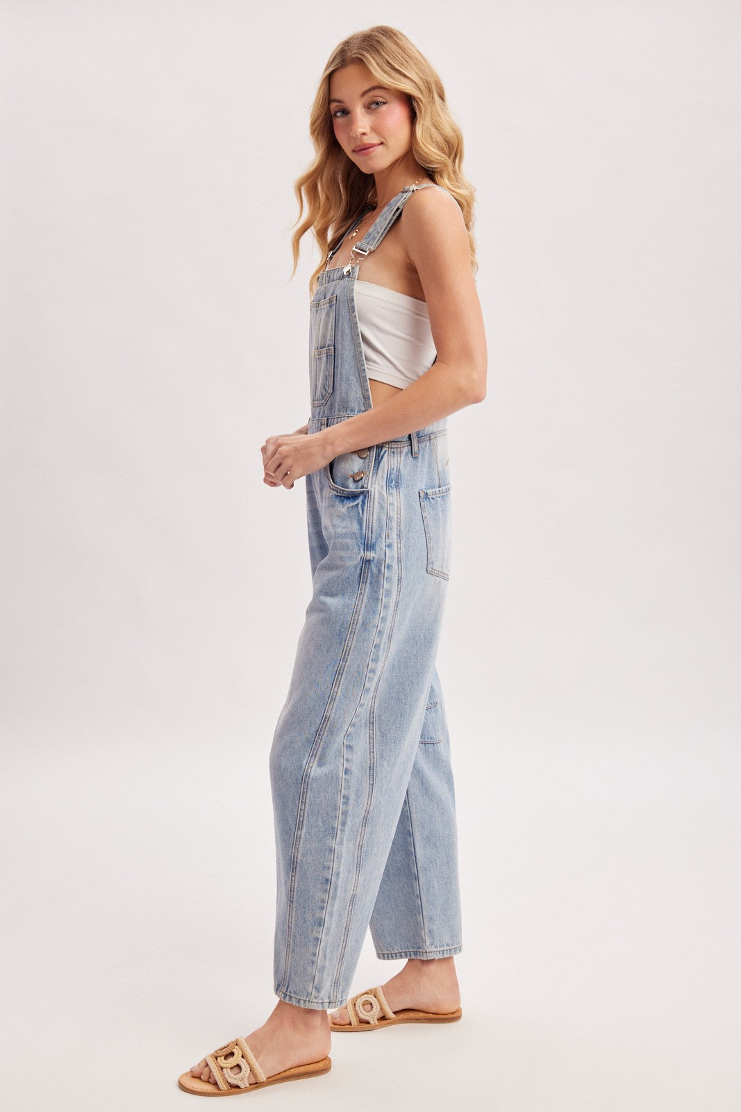 woman wearing light blue denim overalls with a white top, posing sideways, and paired with flat sandals