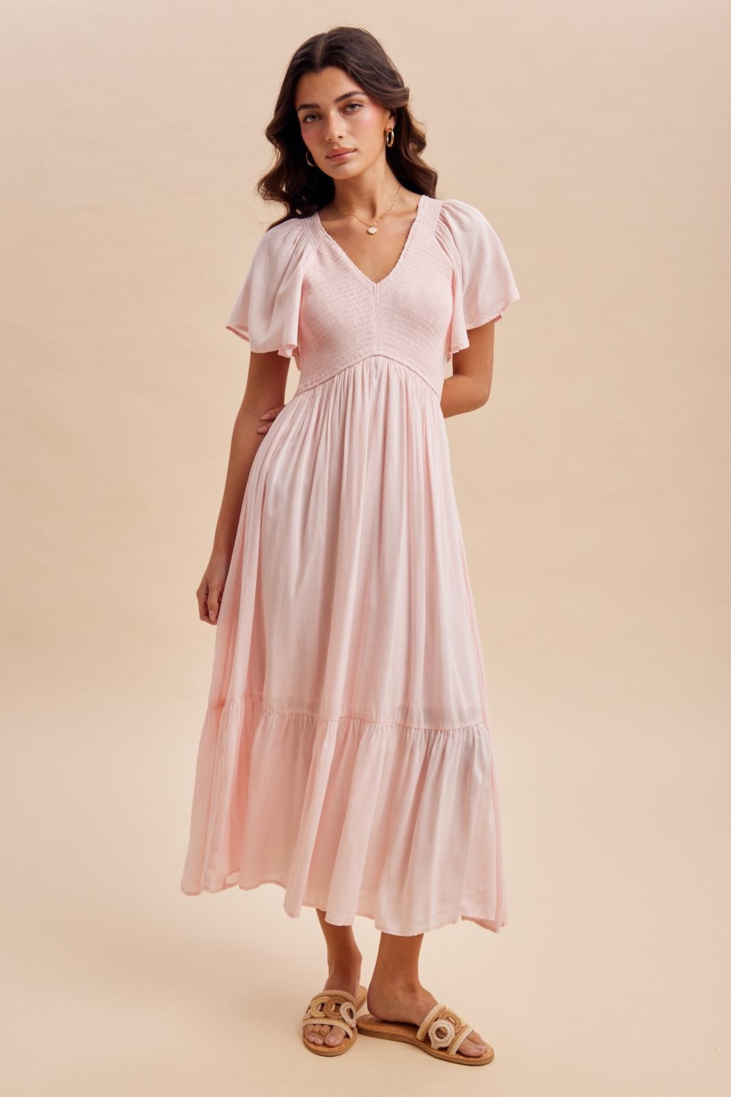 woman wearing a pink maxi dress with short flutter sleeves and a tiered hem, standing against a neutral background