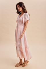 woman wearing a light pink midi dress with flutter sleeves and a tiered skirt, standing sideways on a neutral background