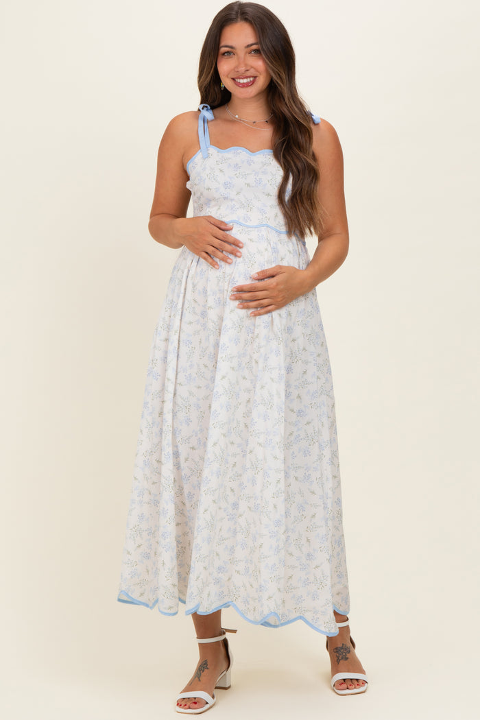 woman wearing a white floral maternity maxi dress with blue accents and a baby bump, standing and smiling