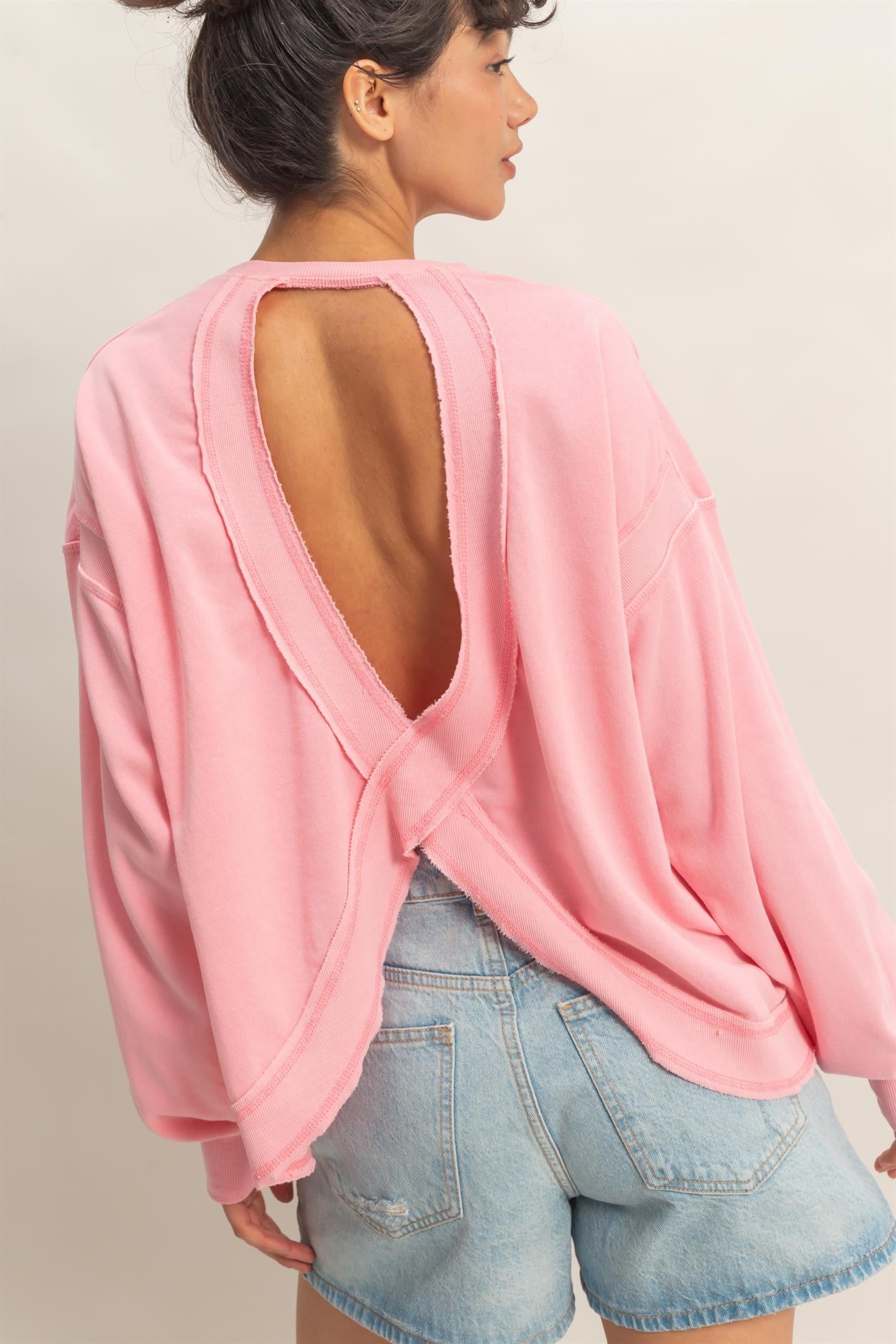 woman wearing a pink oversized pullover sweatshirt with an open back and long sleeves, paired with denim shorts