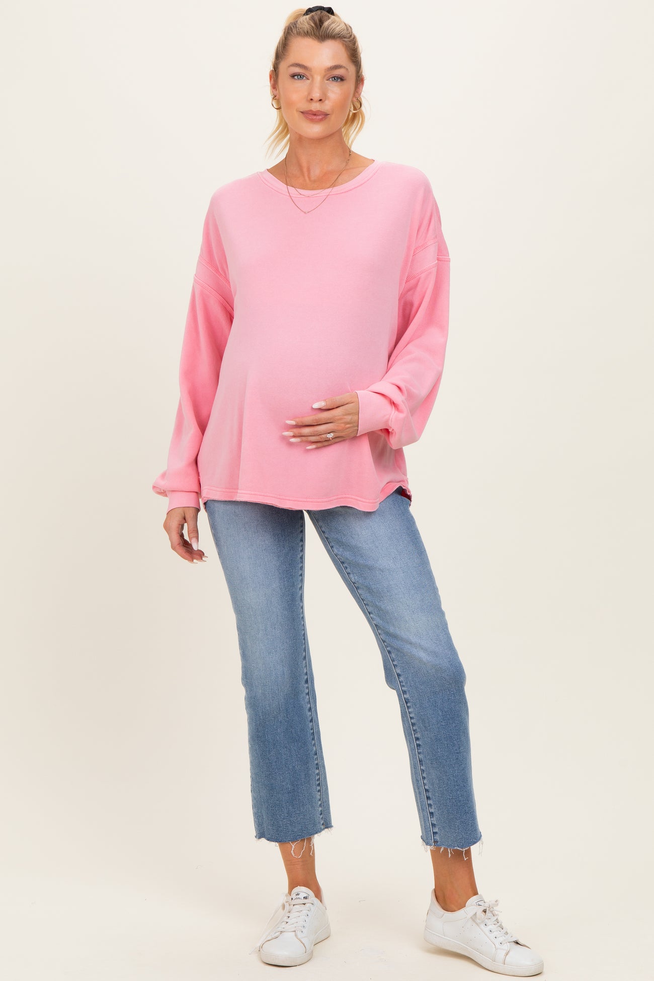 woman wearing a pink oversized maternity top and blue cropped jeans, holding her baby bump