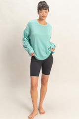 woman wearing a light teal oversized sweatshirt and black bike shorts, standing barefoot against a neutral background