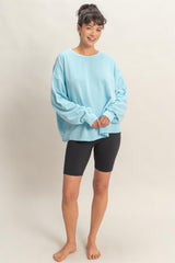 woman wearing a light blue oversized sweatshirt and black biker shorts, standing barefoot and smiling