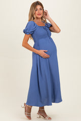 woman wearing a blue maternity maxi dress with puff sleeves and a smocked bodice, holding her baby bump