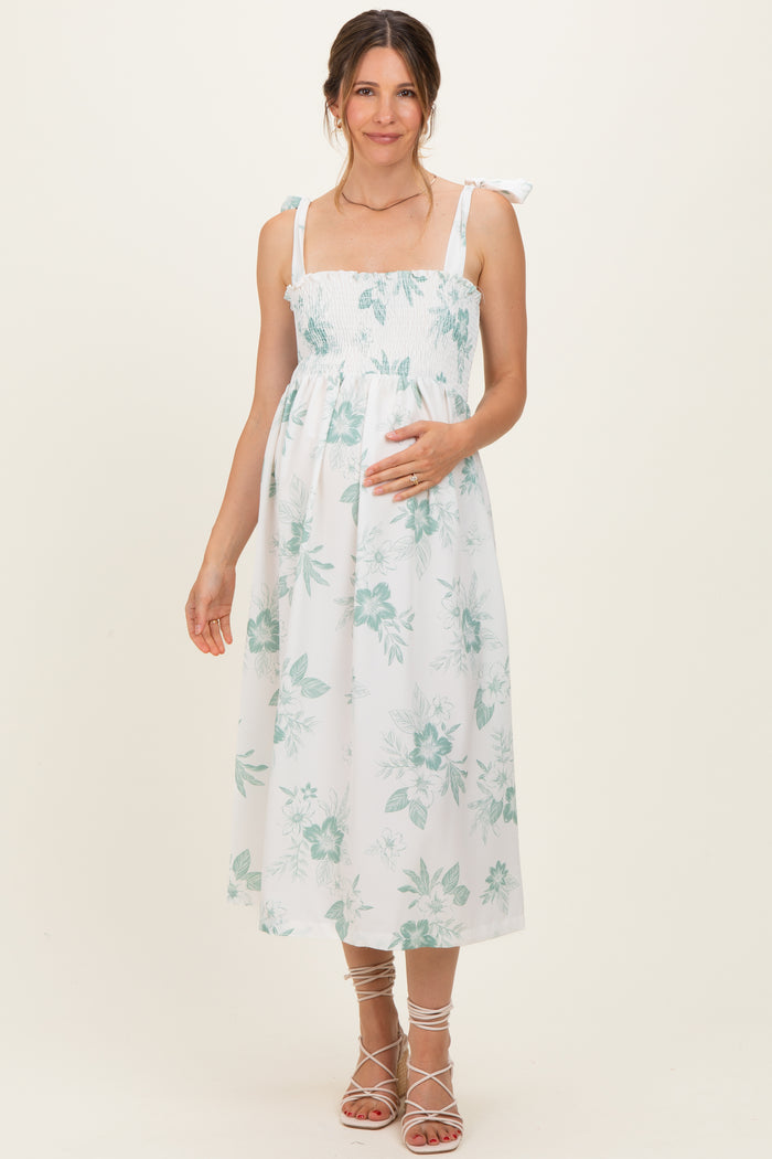 woman wearing a white floral maternity dress with smocked bodice and tie straps, gently holding her baby bump