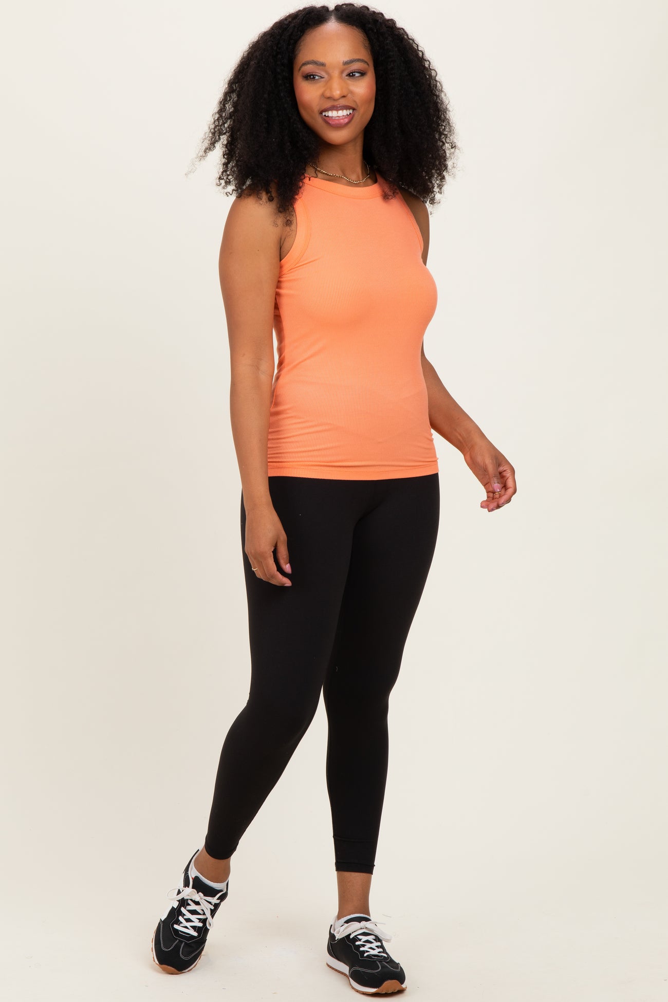 woman wearing an orange sleeveless workout tank top and black leggings, posing with athletic shoes
