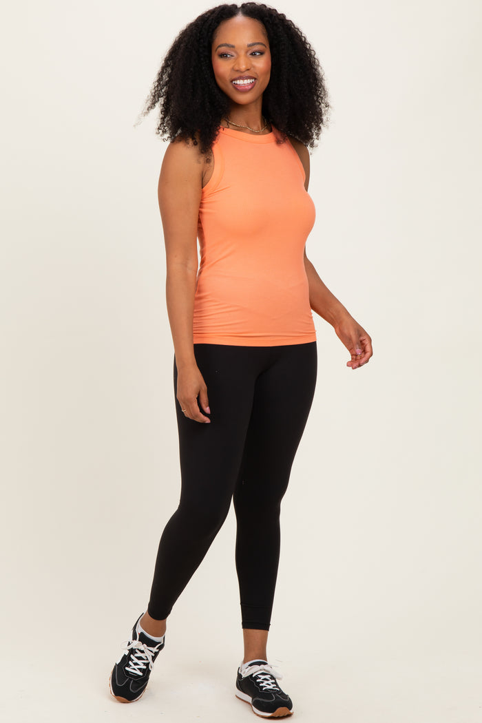woman wearing an orange sleeveless workout tank top and black leggings, posing with athletic shoes