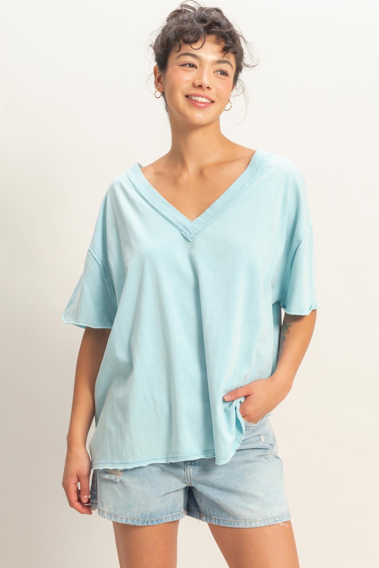woman wearing a light blue oversized v-neck t-shirt and denim shorts, smiling with one hand in her pocket