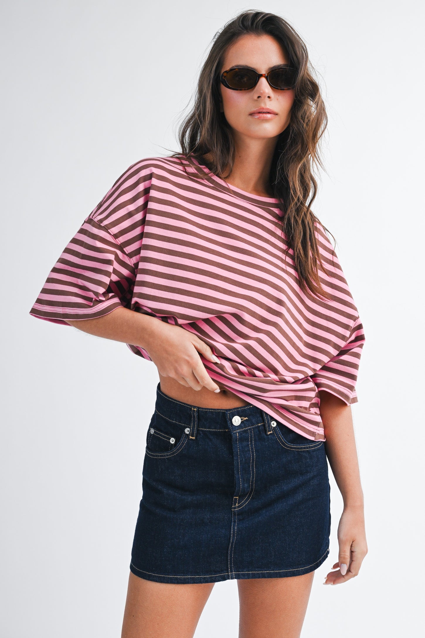 woman wearing a pink and white striped oversized t-shirt and a dark denim mini skirt, styled with sunglasses