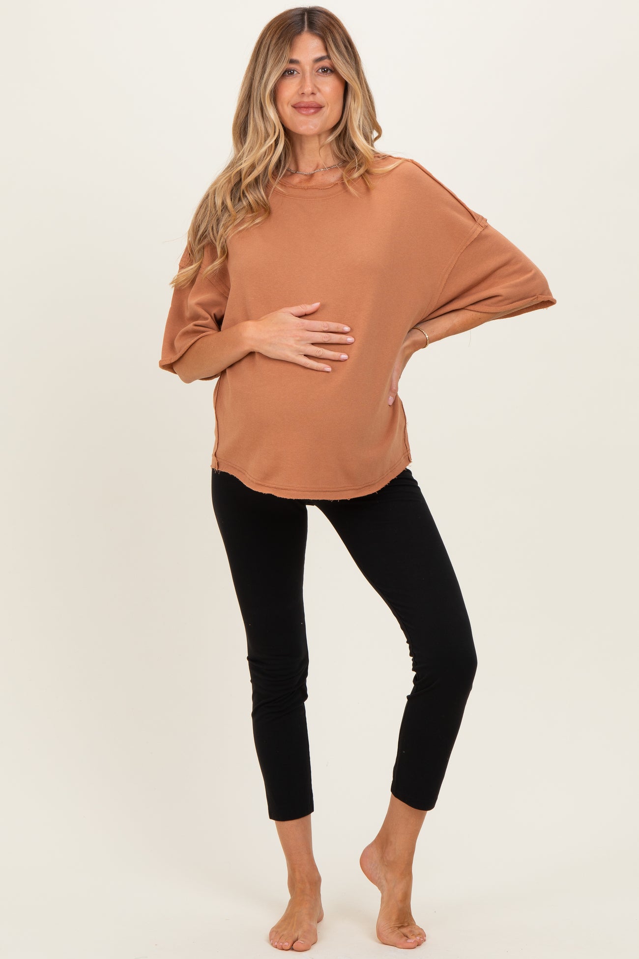 woman wearing a rust colored oversized maternity top and black leggings, holding her baby bump